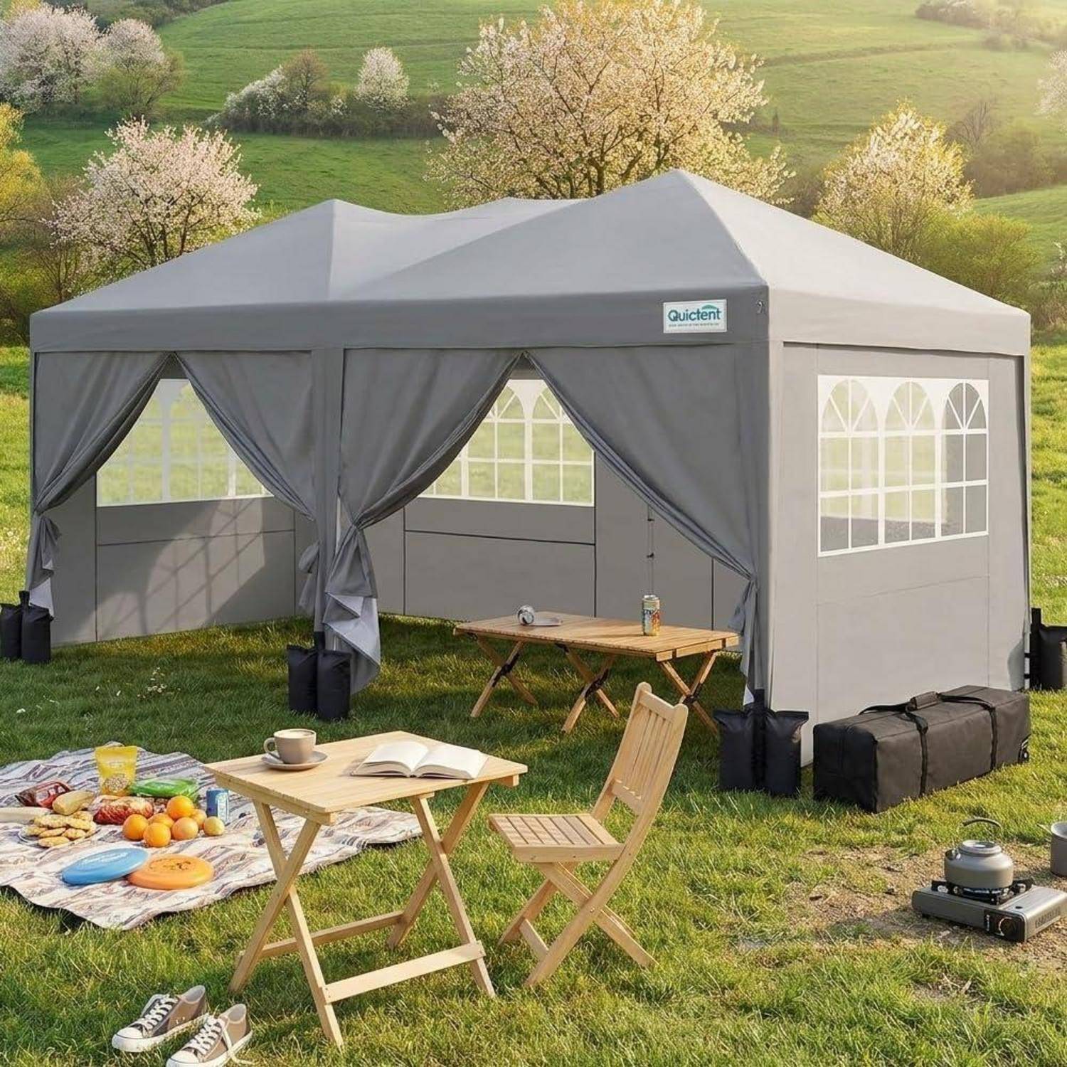 Grauer Pop-Up-Pavillon mit Vorhängen auf einer Wiese, aufgebaut für ein Outdoor-Picknick mit Klapptisch, Stühlen, Essen und Campingausrüstung.
