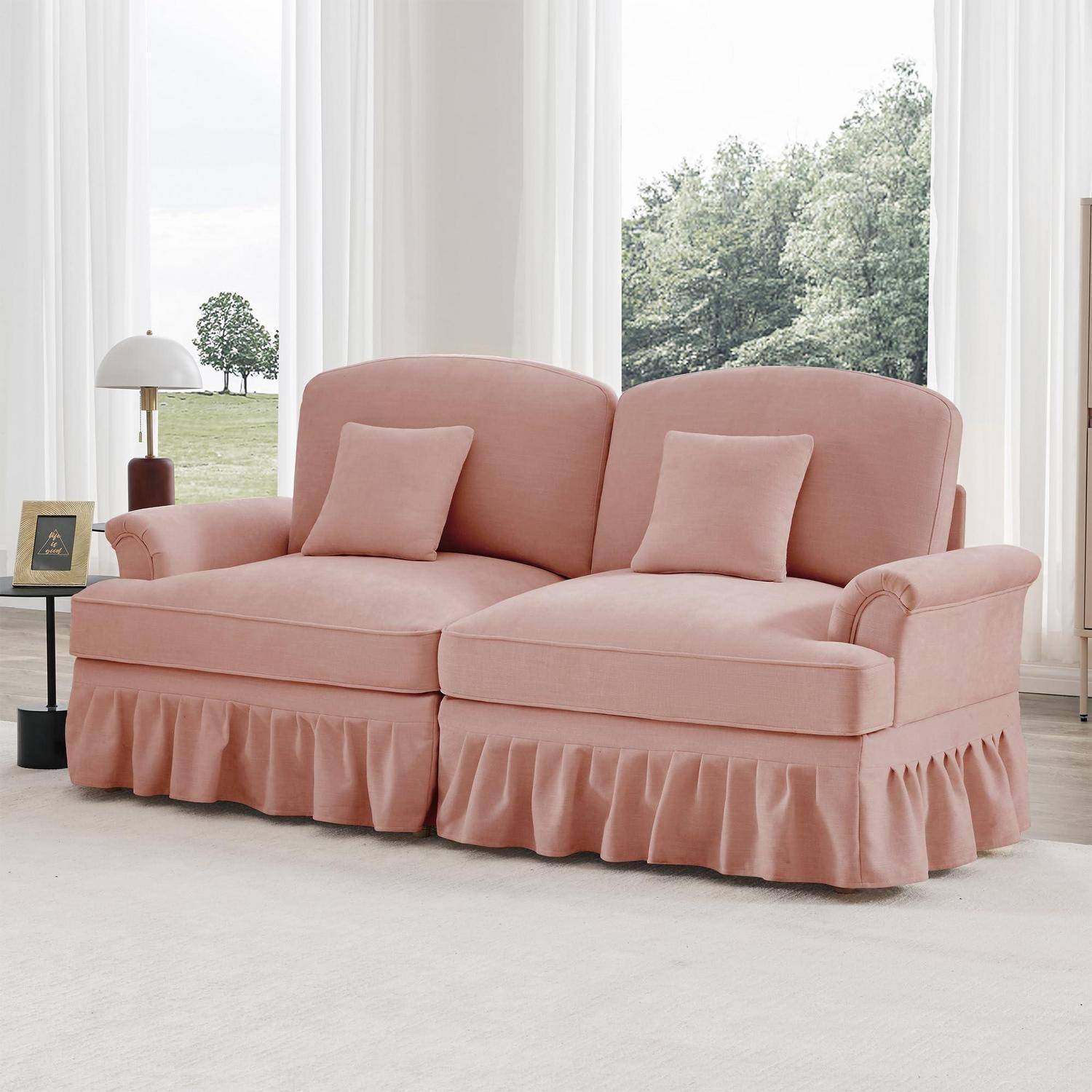 Rosa Zweisitzer-Sofa mit zwei Kissen und einer Sockelbasis in einem hellen Raum mit großen Fenstern.