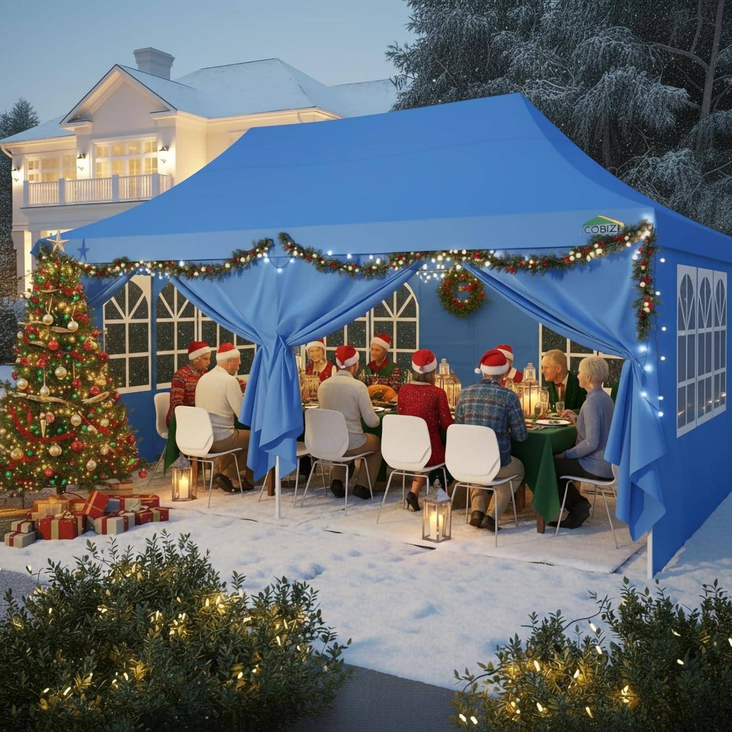 Menschen in Weihnachtsmützen speisen bei einem festlichen Weihnachtsfest in einem blauen Outdoor-Zelt, das mit Lichtern, Girlanden und einem Weihnachtsbaum in schneebedeckter Umgebung dekoriert ist.
