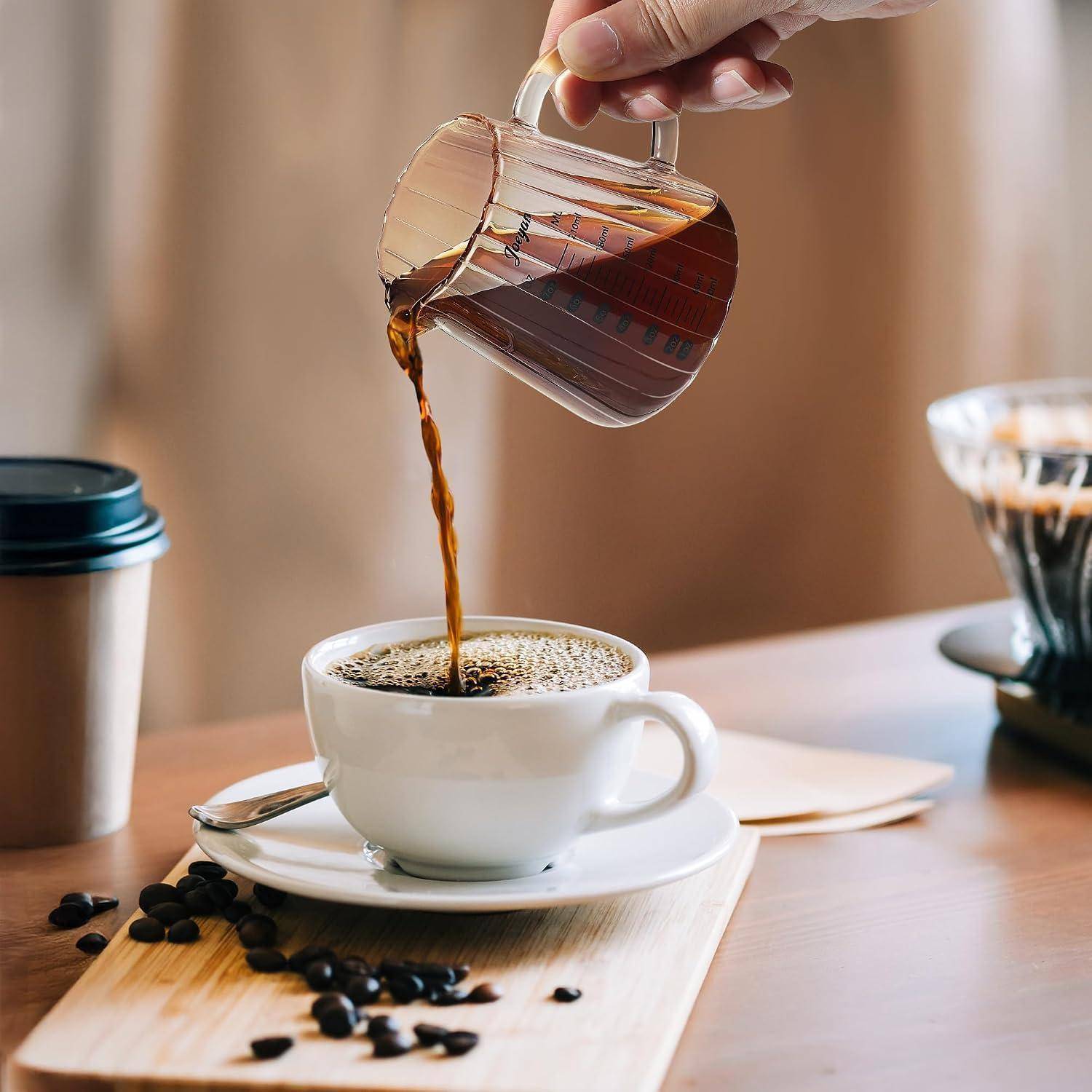 Eine Hand gießt Kaffee aus einem glsernen Dripper in eine weiße Tasse auf einer Untertasse, mit Kaffeebohnen und Brau-Setup auf einem Tisch.
