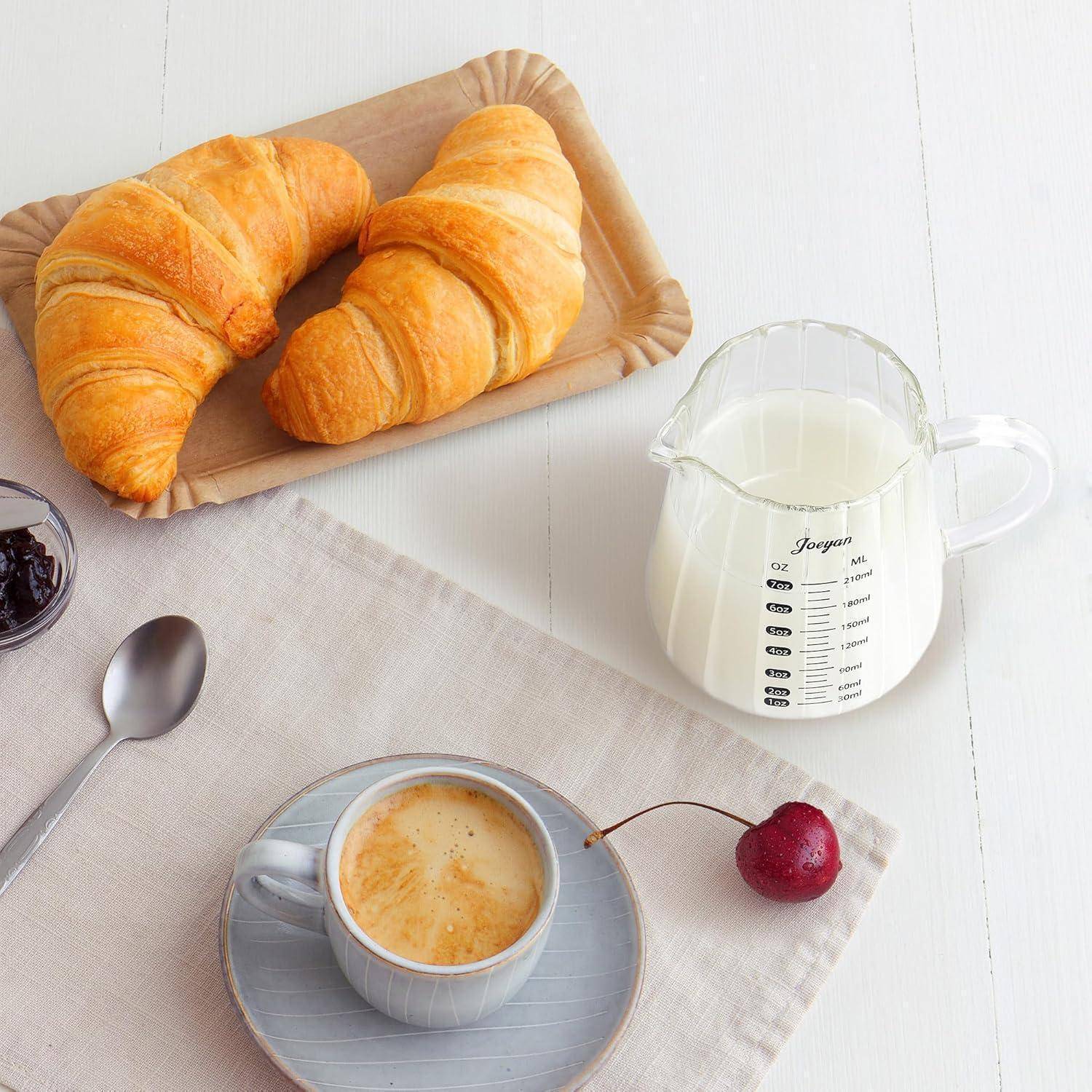 Frühstücksszene mit zwei Croissants auf einem Tablett, einer Tasse Espresso auf einer Untertasse und einem Messbehälter mit Milch auf einem weißen Tisch.