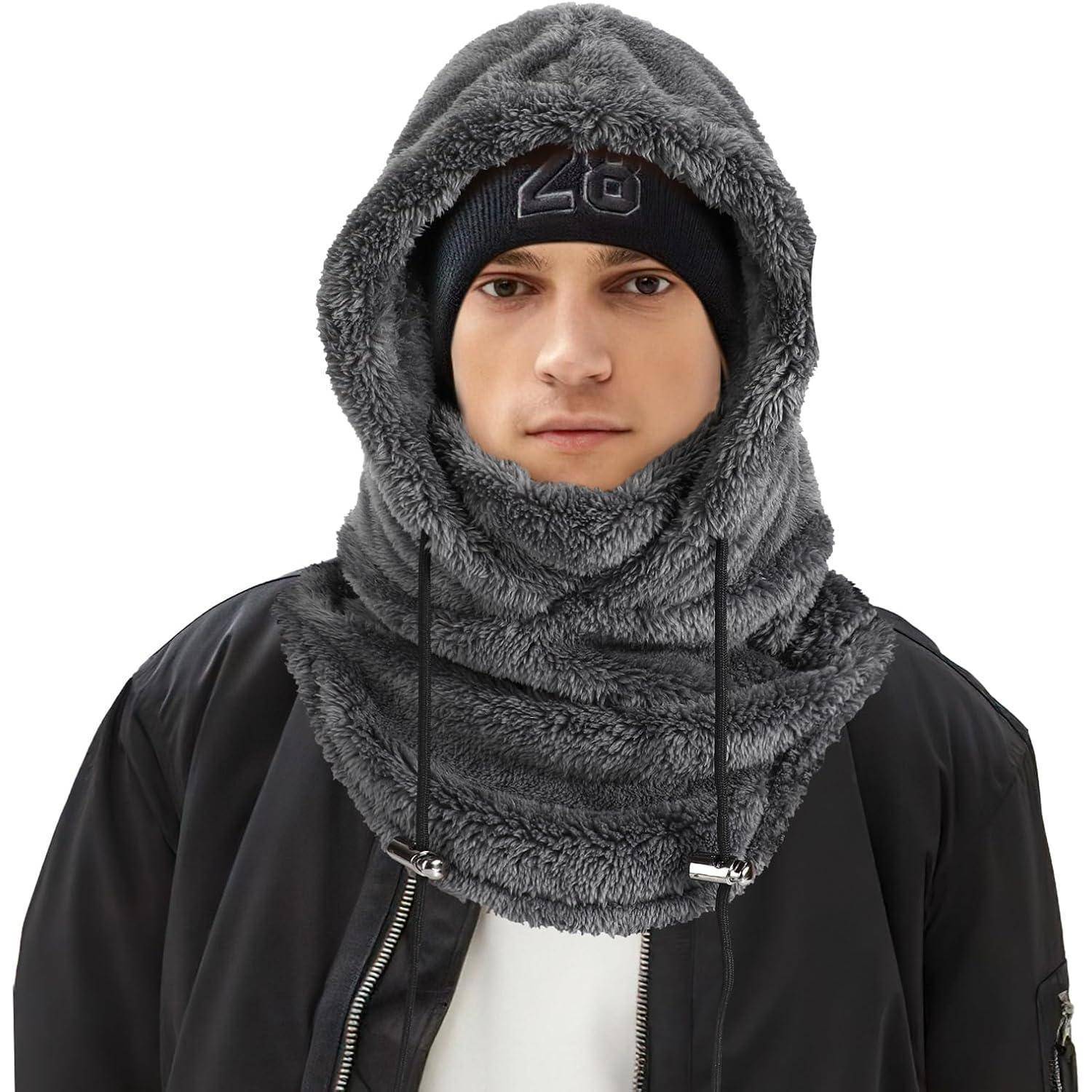 Ein junger Mann trägt eine graue flauschige Skimaske und eine schwarze Mütze, wobei nur sein Gesicht gegen einen einfachen weißen Hintergrund sichtbar ist.
