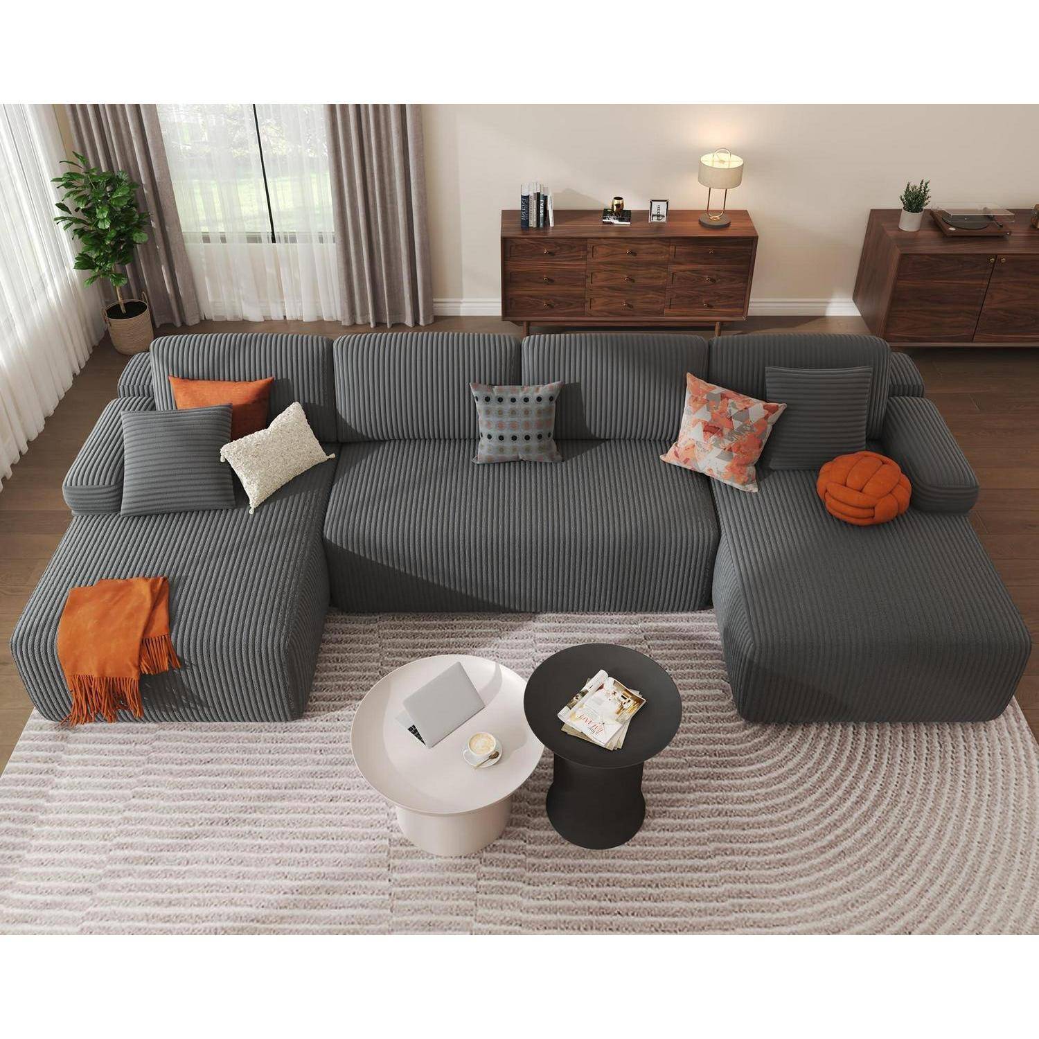 Modernes Wohnzimmer mit großem graugestreiftem Ecksofa, farbenfrohen Kissen, zwei runden Couchtischen und hölzernen Sideboards neben hohen Fenstern.