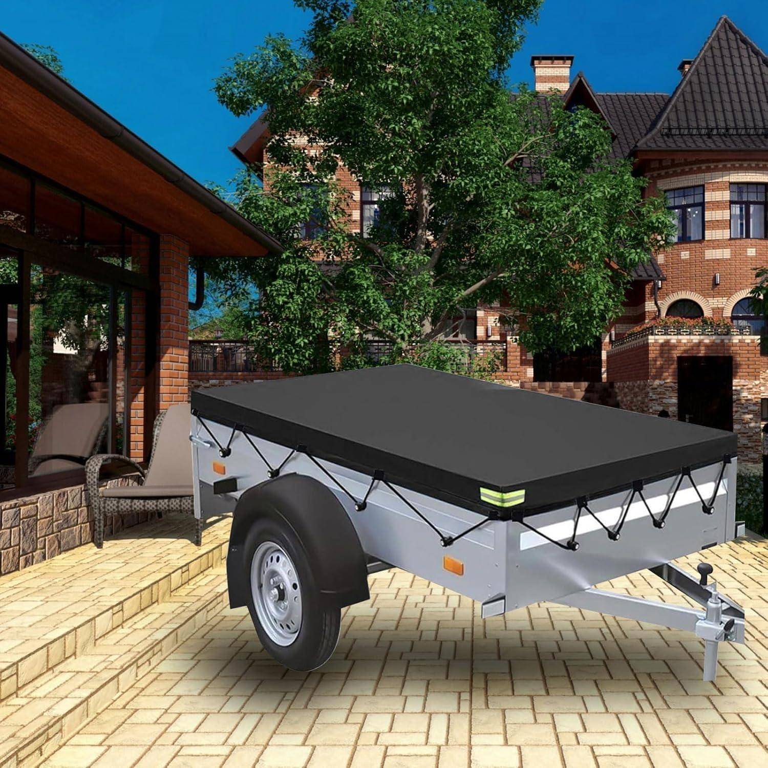Kleiner silberner Mehrzweckhänger mit schwarzer Plane und einem sichtbaren Rad, der auf einer Ziegelpflasterauffahrt neben einem Haus geparkt ist.