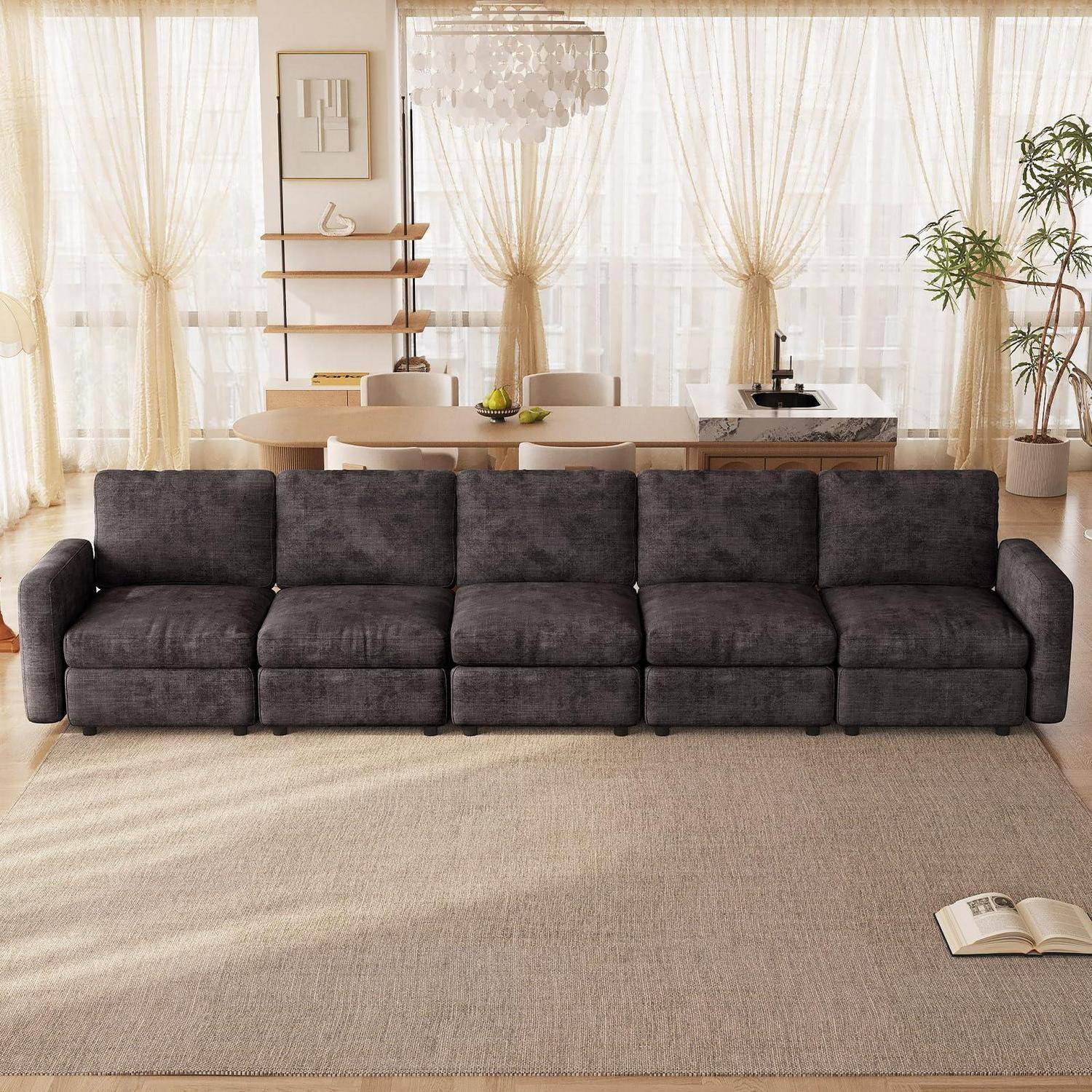 Dunkelgraues modulares Sofa in einem hellen Wohnzimmer mit transparenten Vorhängen, Esstisch, Regalen und einem beigen Teppich mit einem aufgeschlagenen Buch.
