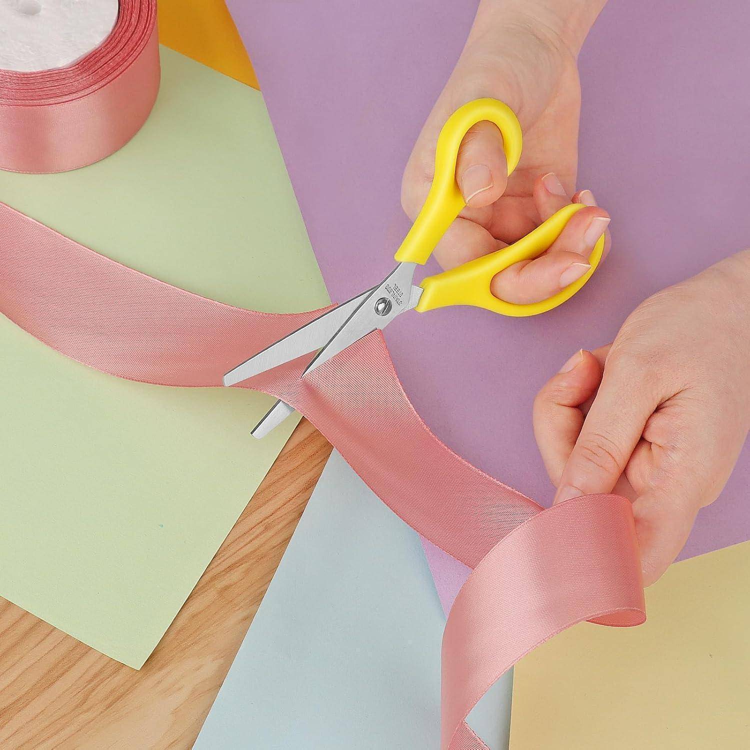 Hände mit gelben Scheren schneiden rosa Schleife über pastellfarbenem Papier auf einer Tischplatte.