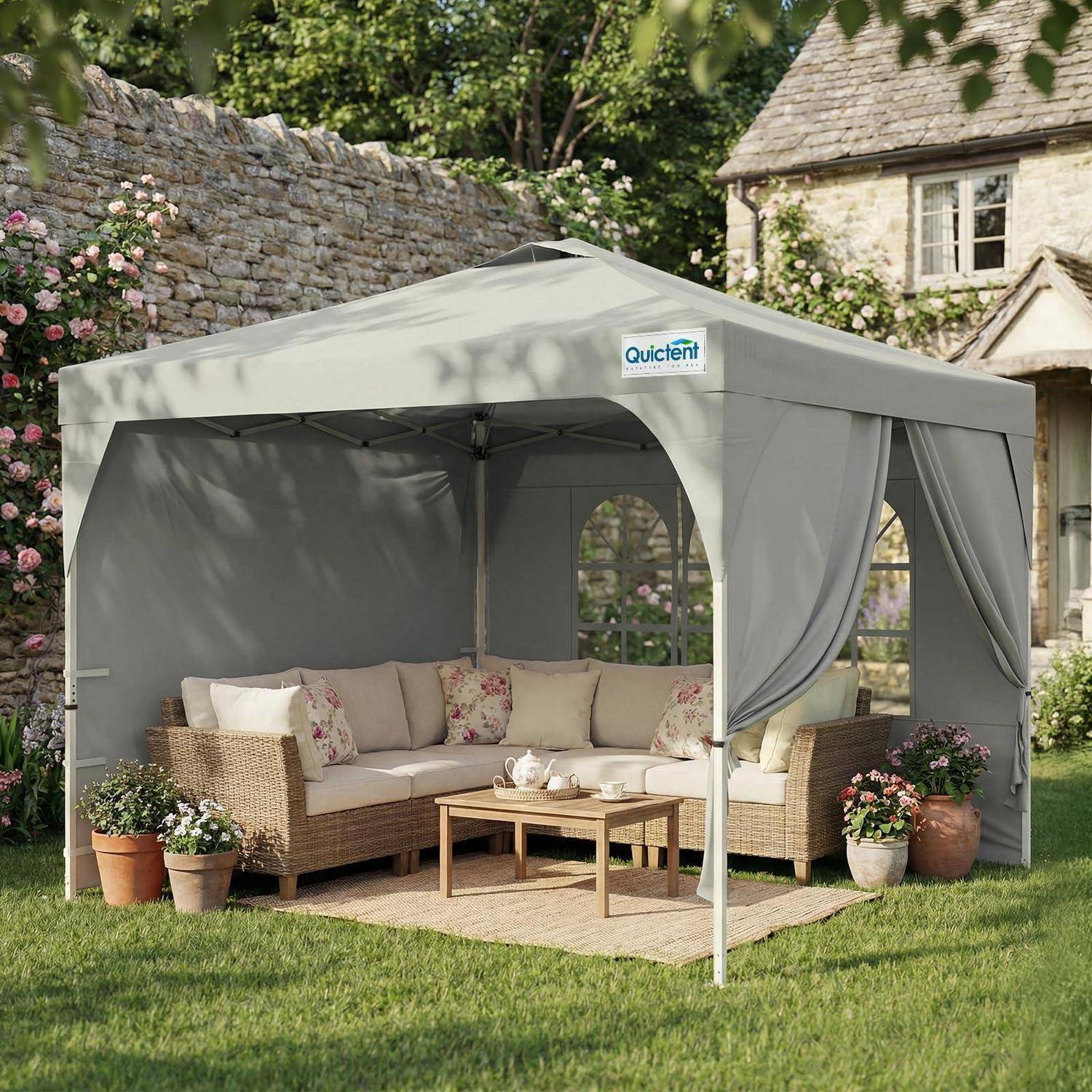 Grauer Gazebo-Baldachin mit Korb-Außensofa, Couchtisch und Topfblumen in einer Gartenumgebung.
