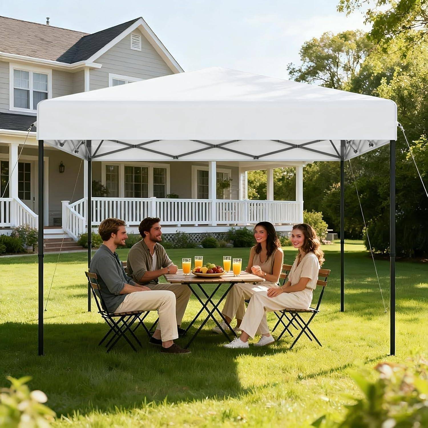 Vier Erwachsene sitzen an einem Klapptisch unter einem weißen Pop-Up-Zelt im Garten eines Vorstadthauses, essen und trinken gemeinsam.