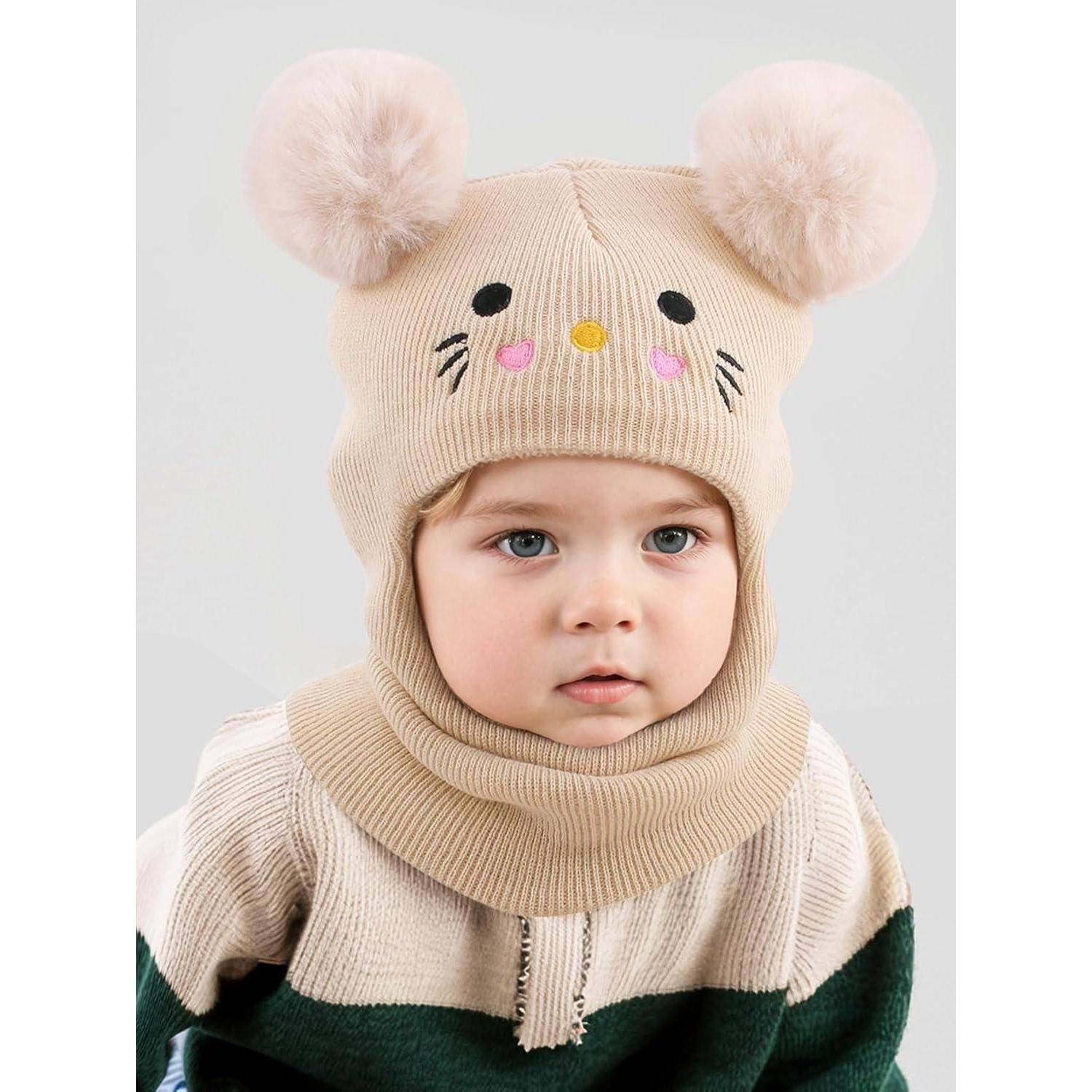 Ein Baby, das eine beige Strickmütze mit Bommel-Ohren und einem Bärgesicht trägt, zusammen mit einem passenden Kapuzenpullover, vor einem einfachen Hintergrund.