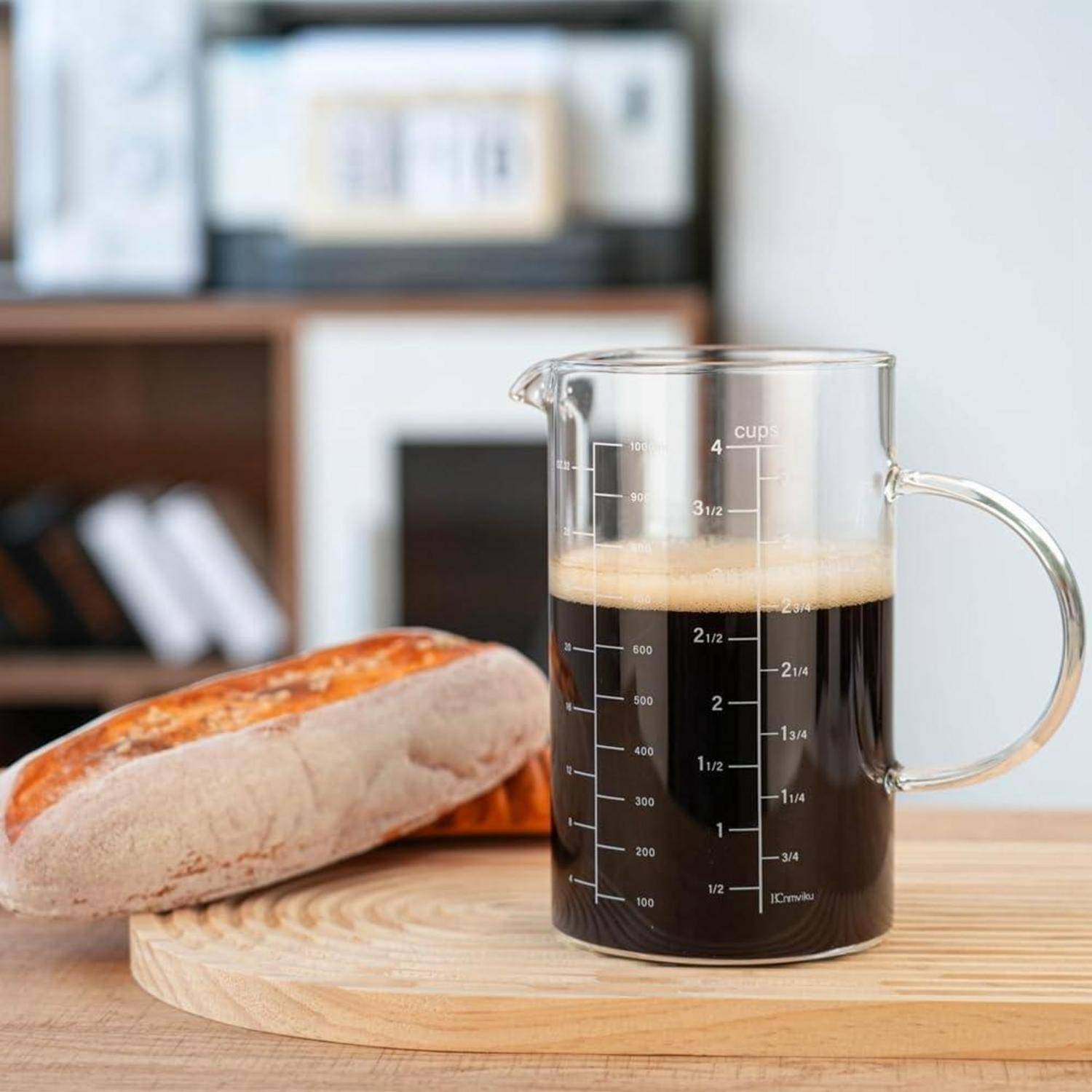 Ein Glaskrug zum Abmessen, gefüllt mit dunklem Kaffee und Schaum, steht auf einem Holzbrett neben zwei Brotscheiben.