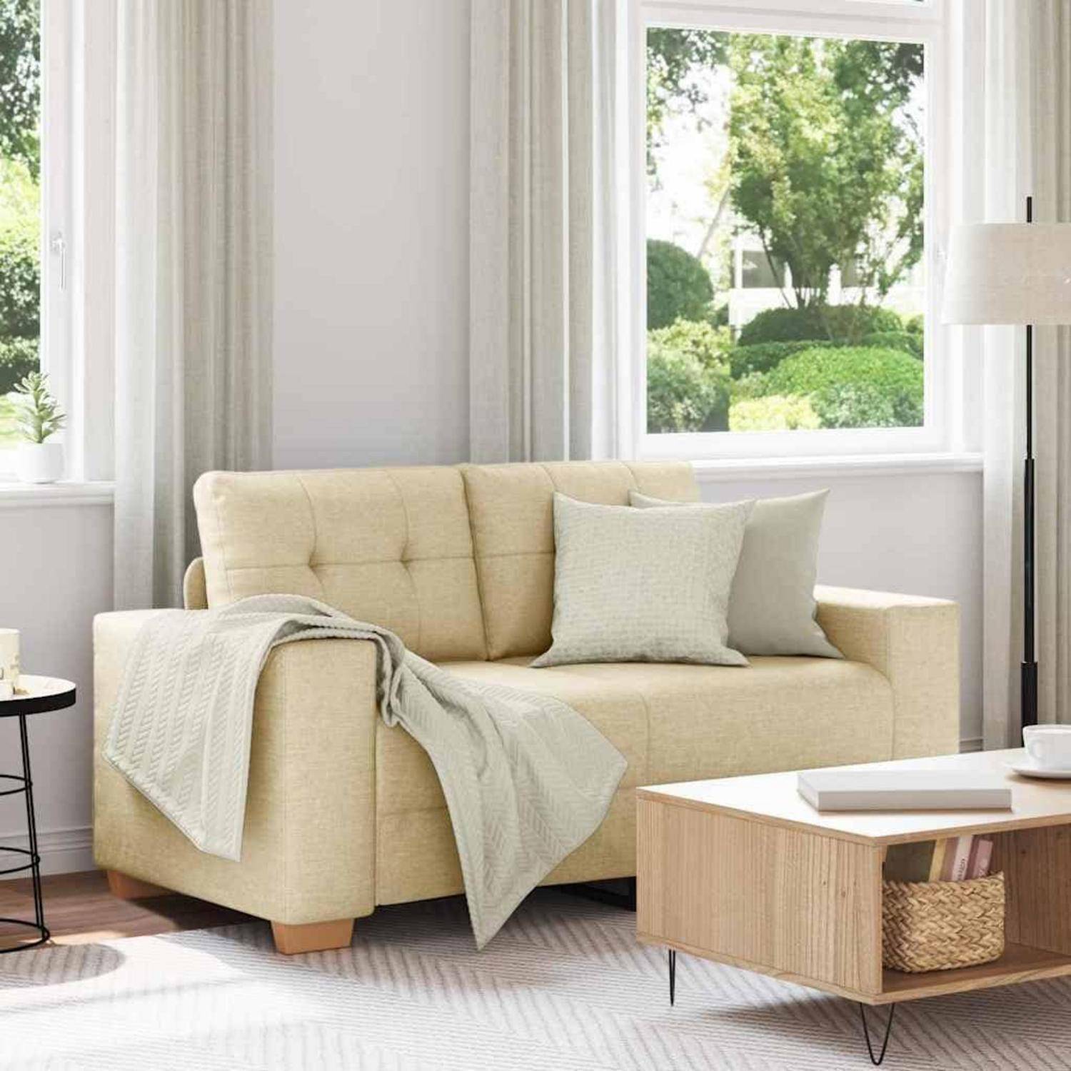 Beige Sofa mit Wurfdecke und Kissen in einem hellen Wohnzimmer neben einem Holz-Couchtisch und großem Fenster mit Blick auf Grünflächen.