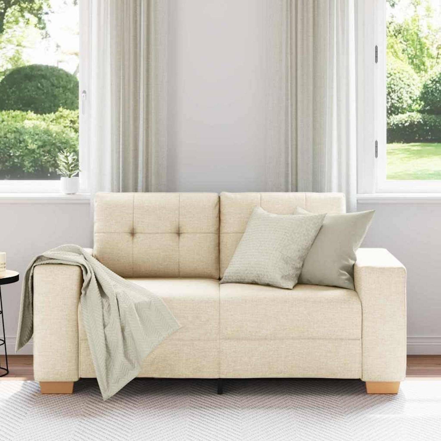 Cremefarbenes Sofa mit zwei Kissen und einer drapierten Wurfdecke in einem hellen Wohnzimmer neben großen Fenstern.
