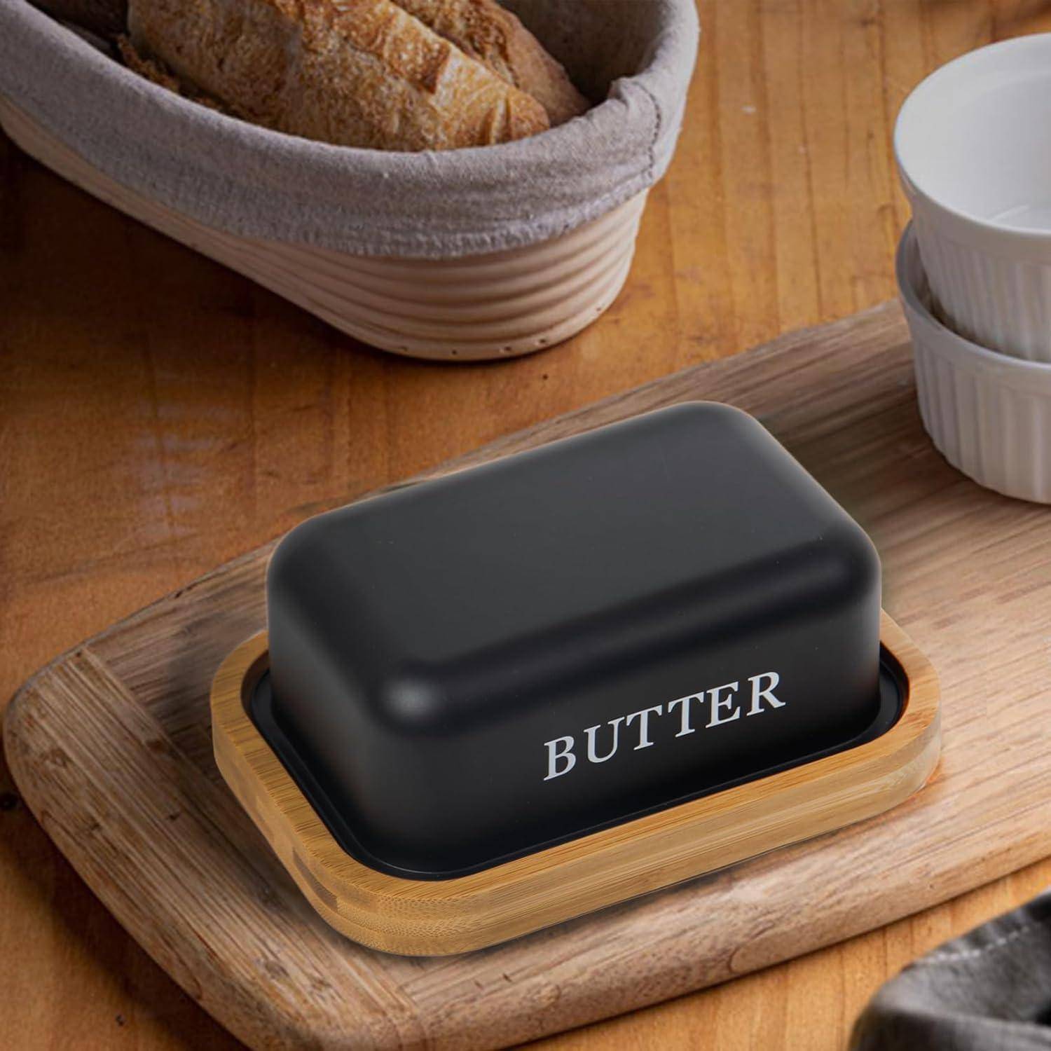 Schwarze Butterdose auf einem Holztablett, mit "BUTTER"" beschriftet, neben Brot und Schüsseln auf einem Küchentisch.