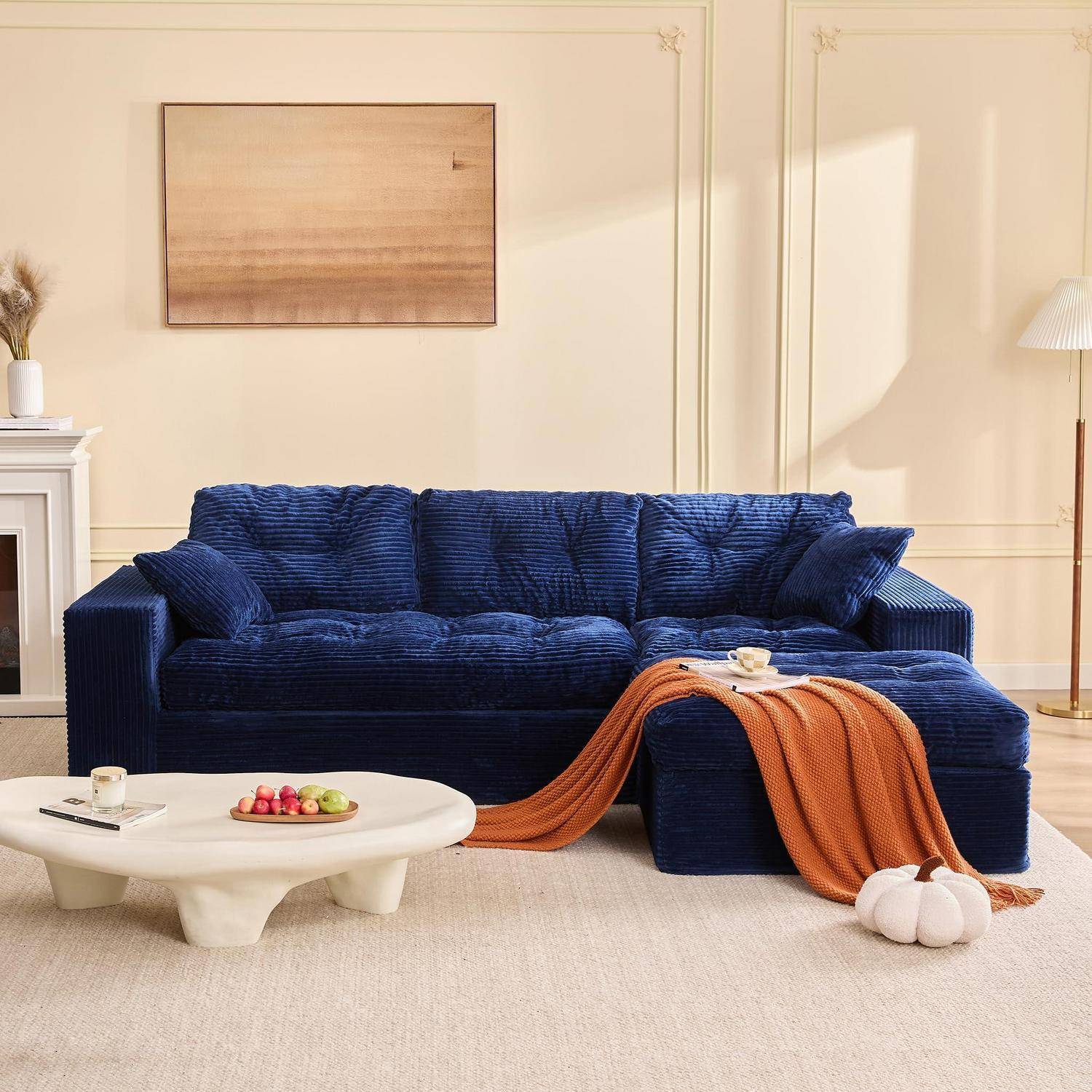 Ein marineblauer Ecksofa mit einer orangefarbenen Wurfdecke in einem hellen Wohnzimmer, neben einem niedrigen weißen Couchtisch mit Obst und Kerzen.
