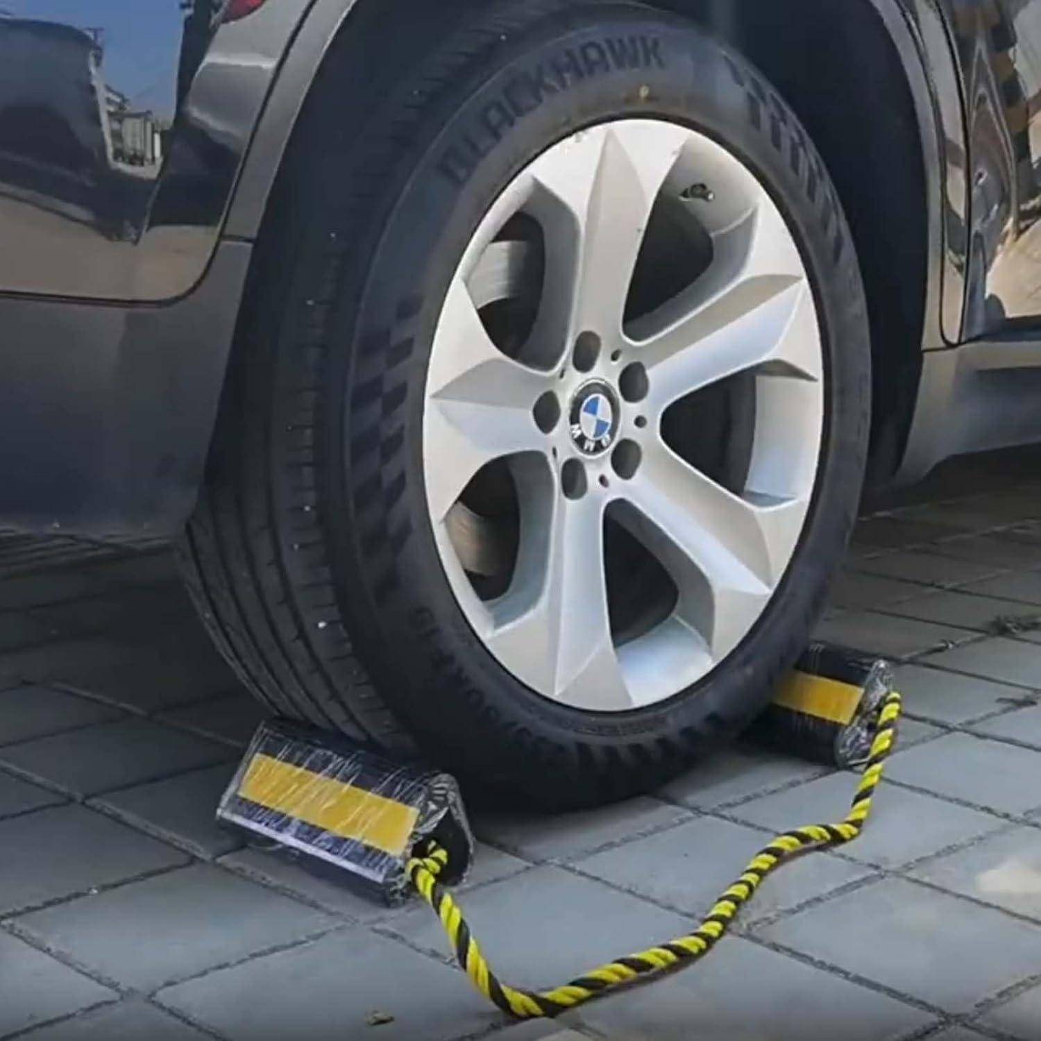 Ein Autoreifen auf einer Einfahrt wird durch zwei gelb-schwarze Radkeile blockiert, die durch ein Seil verbunden sind und eine Bewegung verhindern.