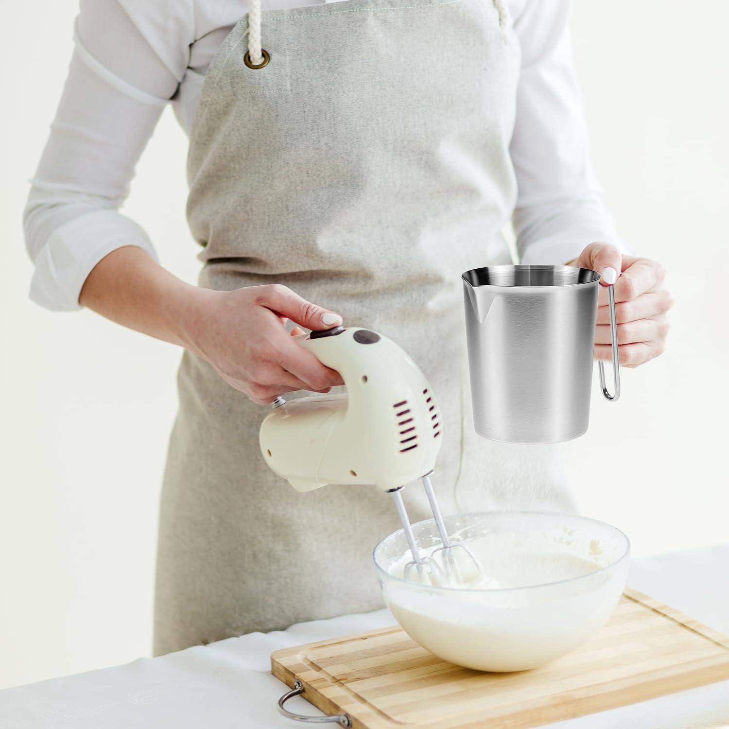 Eine Person in einer Schürze verwendet einen elektrischen Handmixer, um Zutaten in einer Schüssel zu schlagen, während sie eine Metallmesskanne über einem Schneidebrett hält.