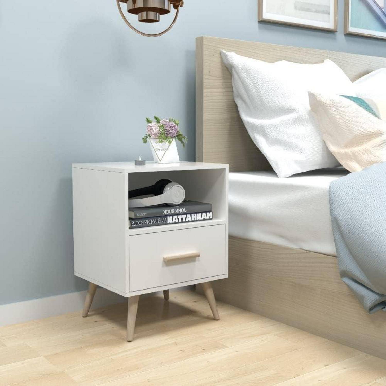 Weißer Nachttisch mit einer Schublade und offenem Regal neben einem Bett, auf dem eine kleine Blumenvase und Bücher in einem hellen Schlafzimmer stehen.