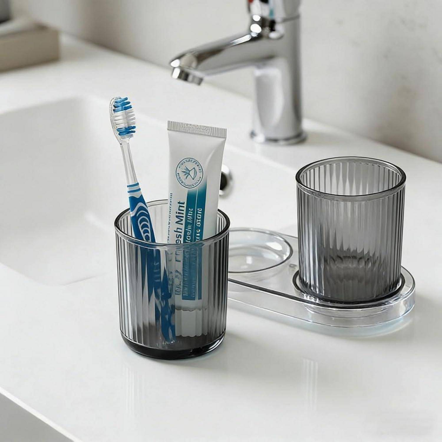 Zahnbürste und Zahnpasta in einem rauchgrauen Badezimmerglas auf einem weißen Waschbecken neben einem passenden leeren Behälter und einem Wasserhahn.