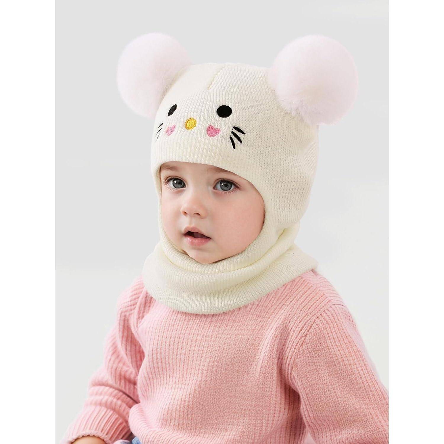 Baby in einem rosa Pullover und einer cremefarbenen Strickmütze mit Mausohren, zur Kamera gewandt vor einem einfachen hellen Hintergrund.