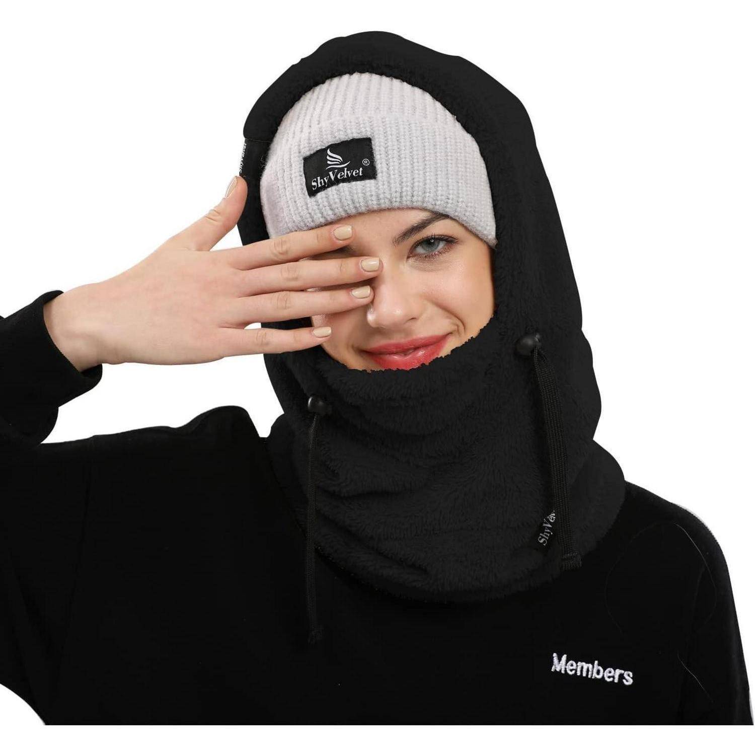 Person trägt eine schwarze Sturmhaube und eine graue Mütze, mit einer Hand, die einen Teil ihres Gesichts verdeckt, auf weißem Hintergrund.