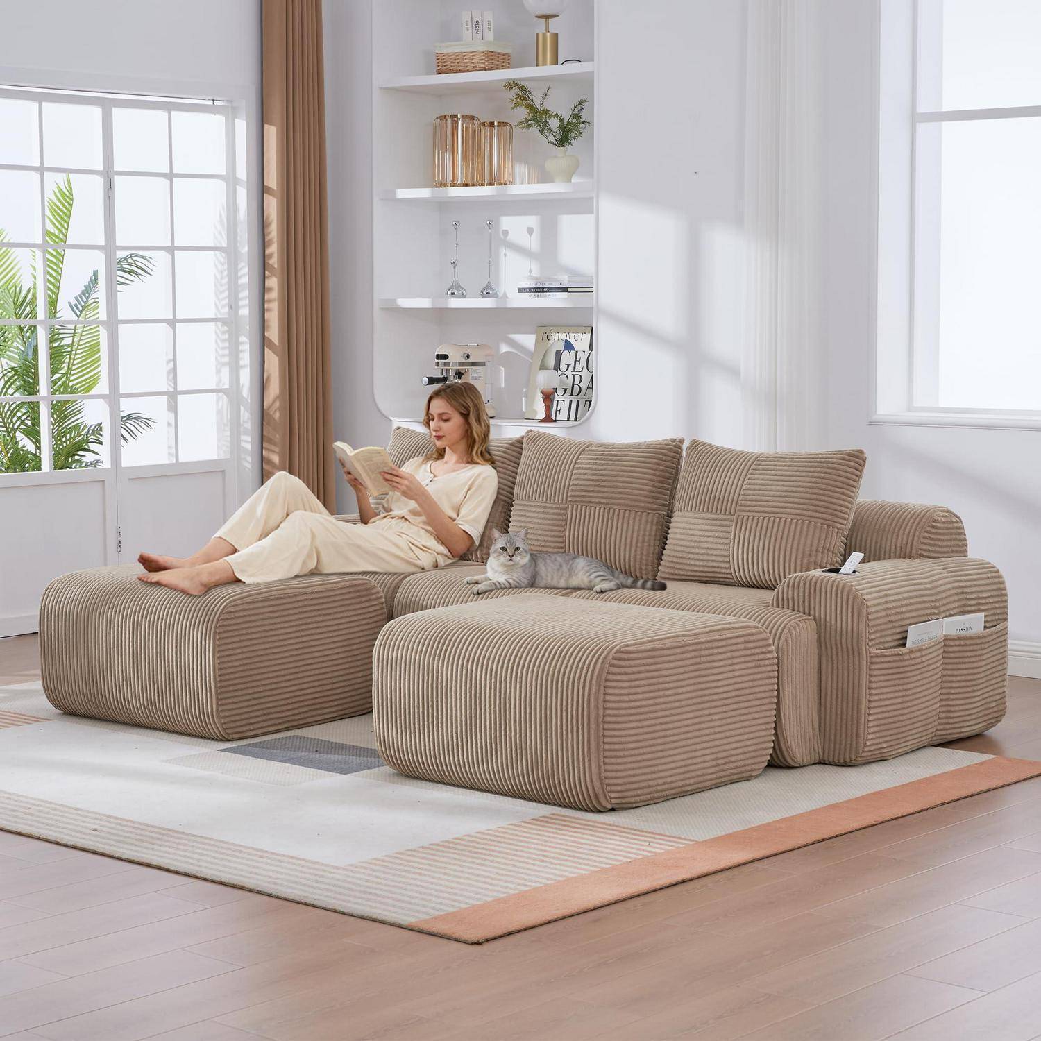 Frau liest auf einem großen beige gestreiften Ecksofa mit einer Katze in einem hellen Wohnzimmer mit weißen Regalen und Fenstern.