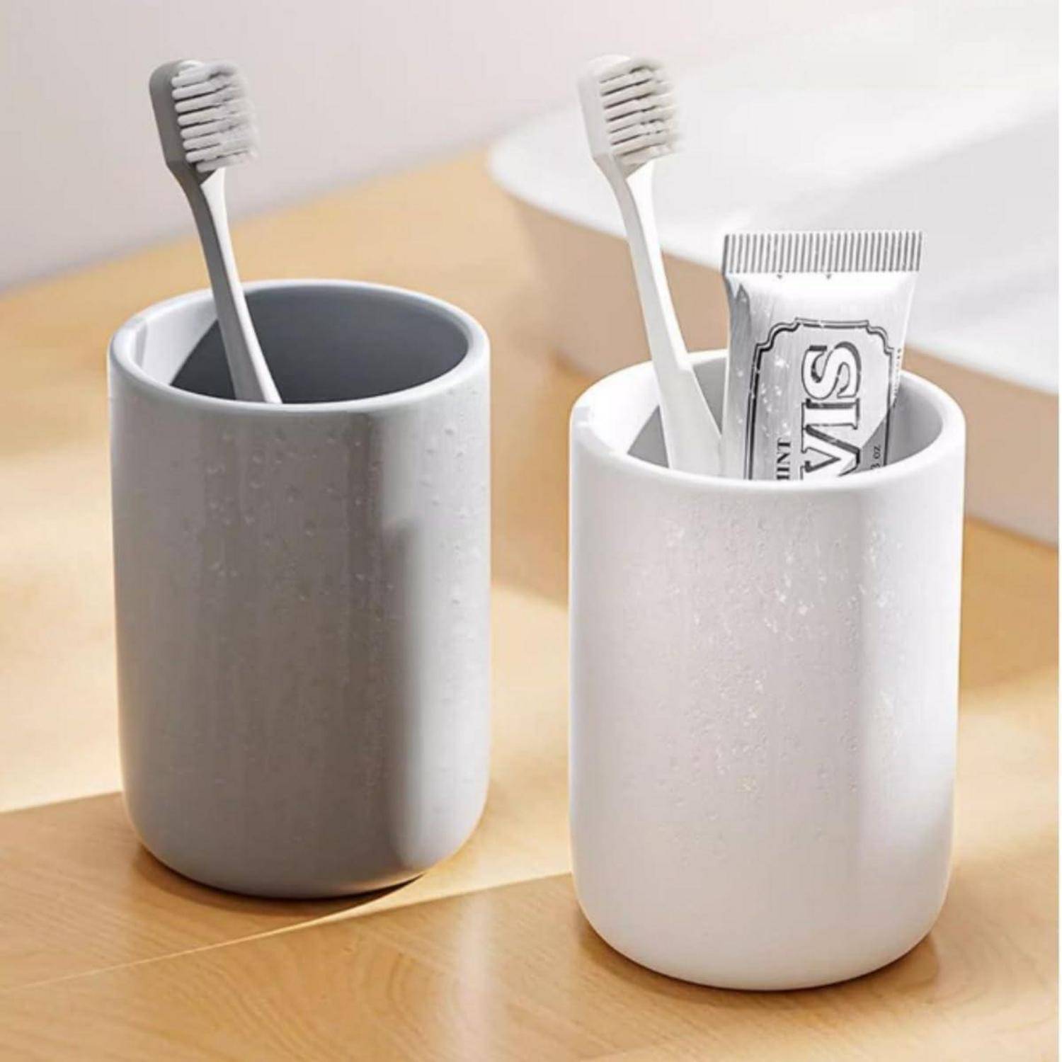 Zwei keramische Badezimmertassen halten Zahnbürsten; die weiße Tasse enthält auch eine Zahnpastatube und zeigt einen einfachen Organizer für die Spüle.