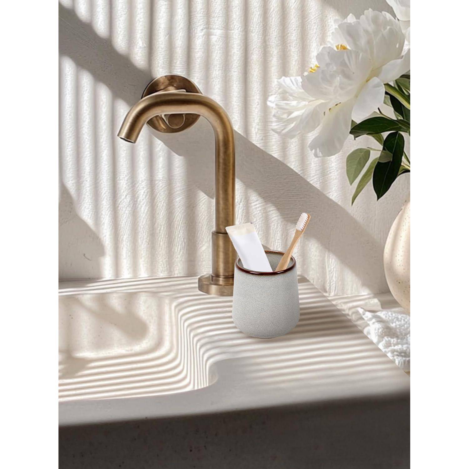 Moderner Messingwasserhahn über einem weißen Waschbecken mit einem Zahnbürstenbecher und Blumen, beleuchtet durch gestreiftes Sonnenlicht und Schatten.