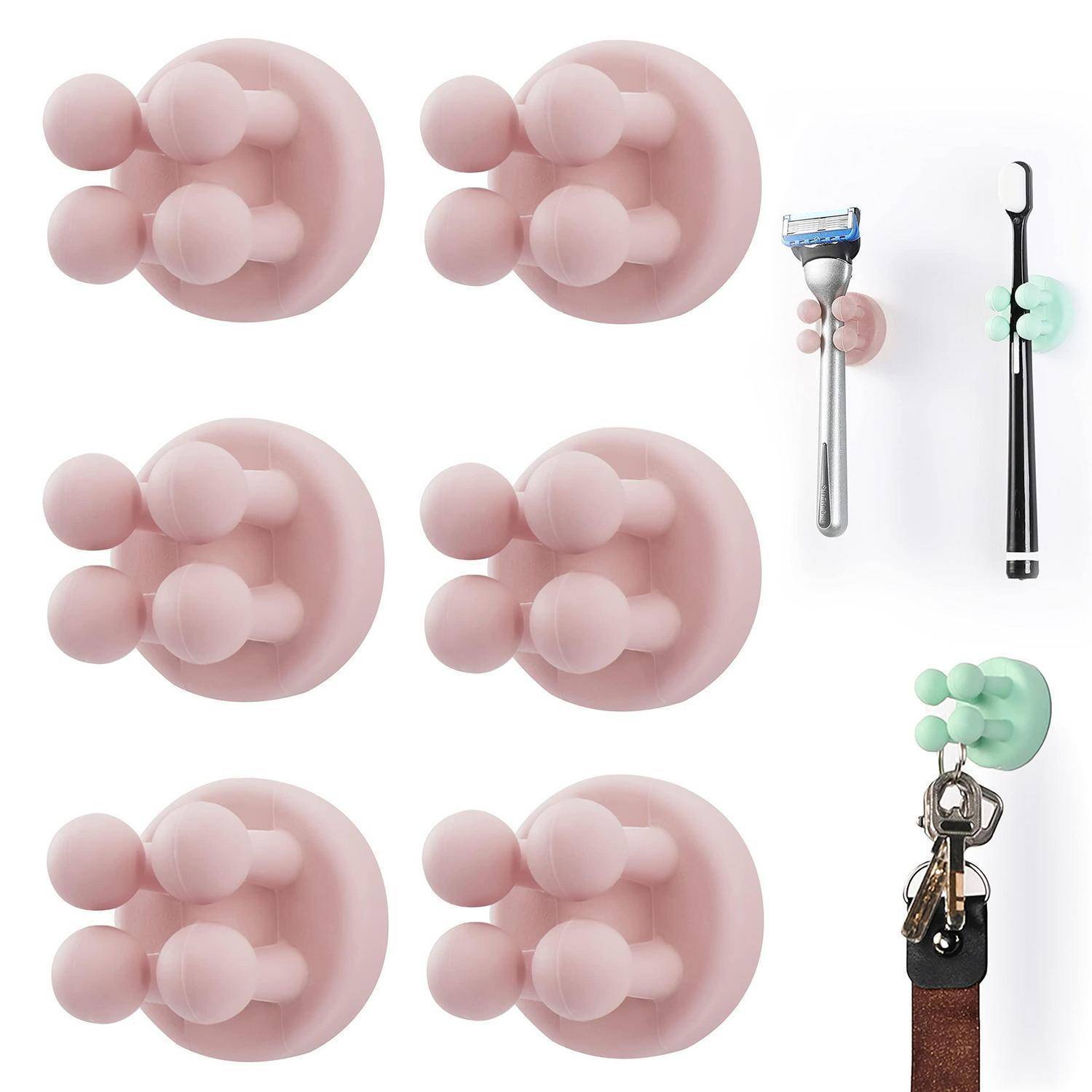 Mehrere haftende Wandhaken in Rosa und Minzgrün halten ein Rasiermesser, eine Zahnbürste und Schlüssel und zeigen verschiedene Befestigungsmöglichkeiten.