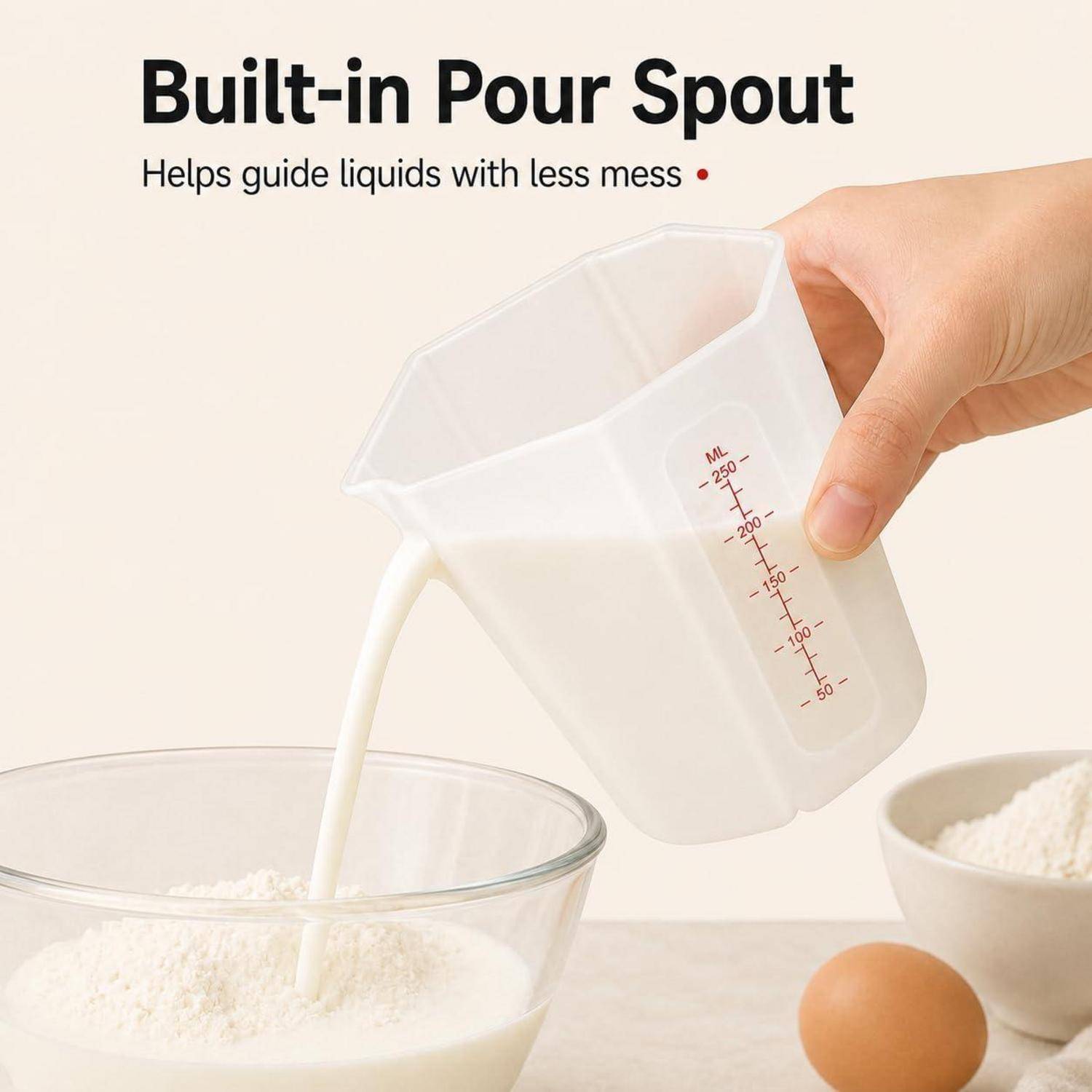 Hand gießt Milch aus einem Messbecher mit integriertem Ausguss in eine Schüssel mit Mehl; der Text lautet „Integrierter Ausguss" und „Hilft, Flüssigkeiten mit weniger Verschmutzung zu führen"".