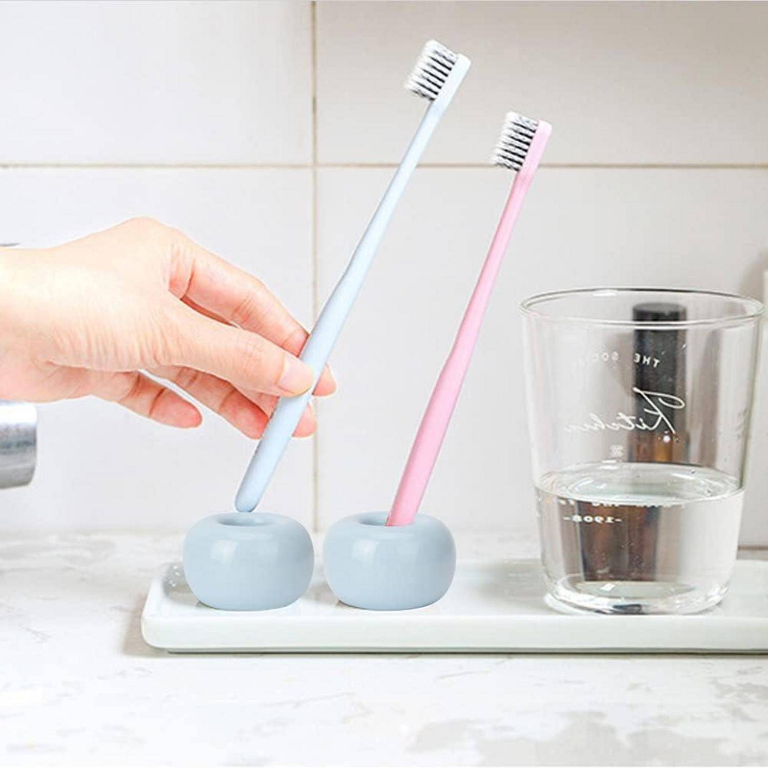 Hand, die eine hellblaue Zahnbürste in einen passenden Halter neben einer rosafarbenen Zahnbürste und einem Glas Wasser auf einem Badezimmertablett legt.