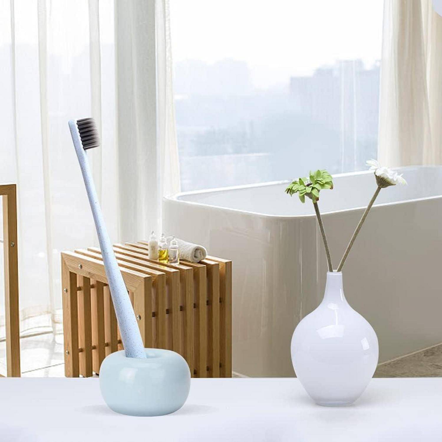 Eine pastellblaue Toilettenbürste in einem abgerundeten Halter steht neben einer weißen Vase mit Blumen in einem hellen, modernen Badezimmer.