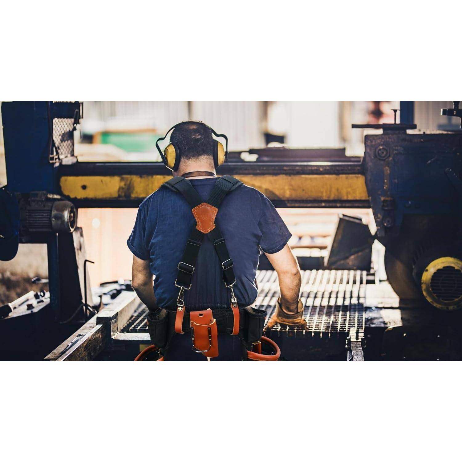 Rückansicht eines Fabrikarbeiters mit Gehörschutz und Sicherheitsgurt, der Industriemaschinen an einer Produktionslinie bedient.