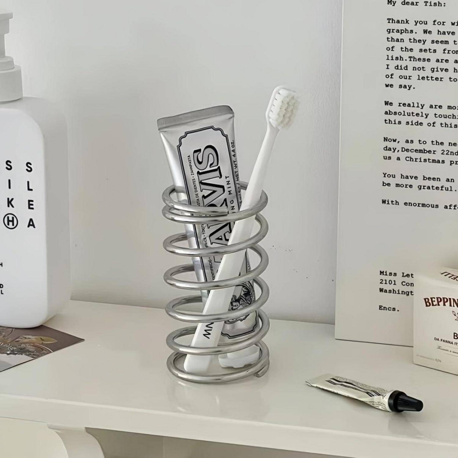 Zahnbürste und Zahnpasta in einem silbernen Spiralhalter auf einem weißen Regal, neben einer weißen Flasche und einer gedruckten Karte.