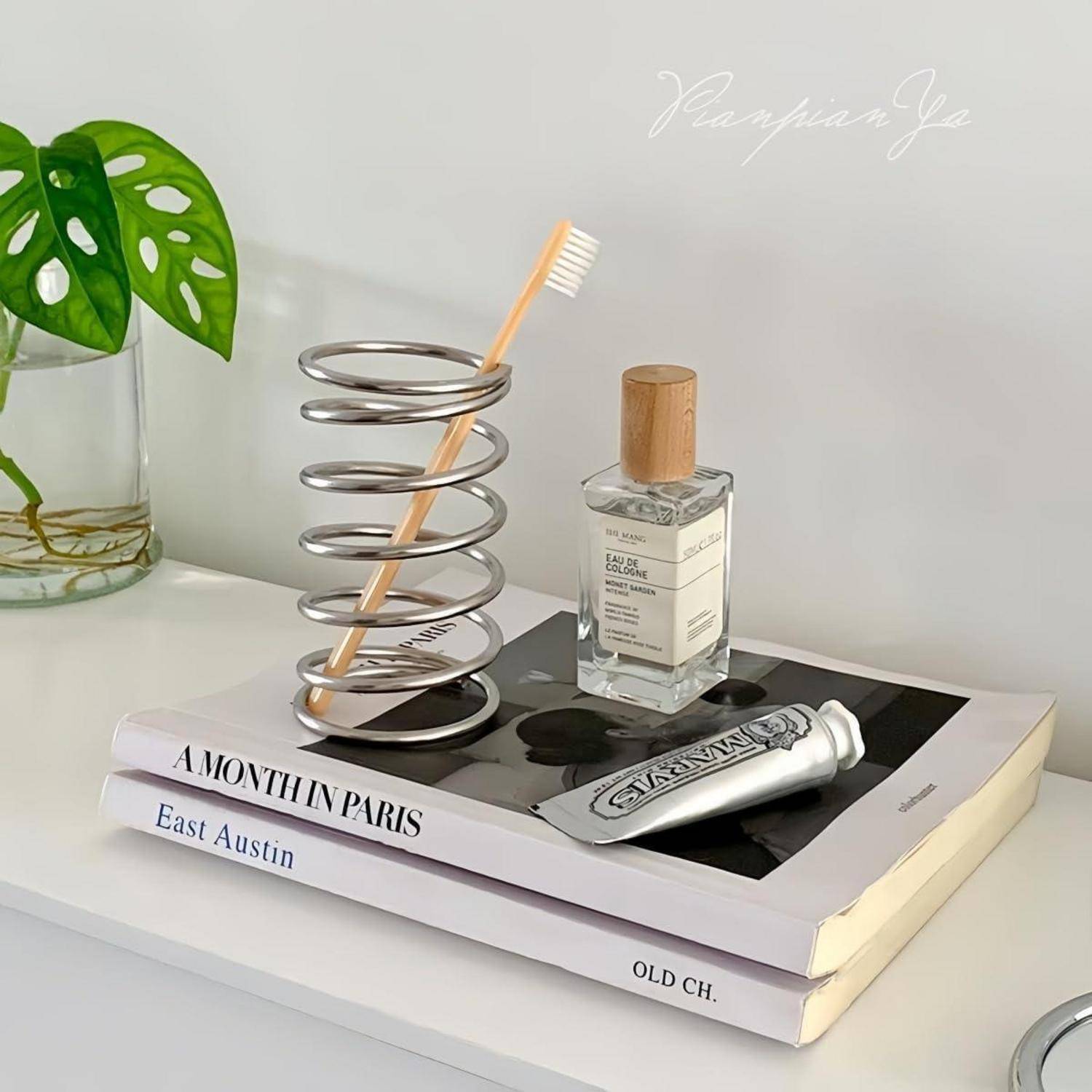 Dekoratives Regalvignette mit gestapelten Büchern, einem spiralförmigen Zahnbürstenhalter, einer Parfümflasche und einer Zahnpastatube neben einer Glasvase mit einem grünen Blatt.