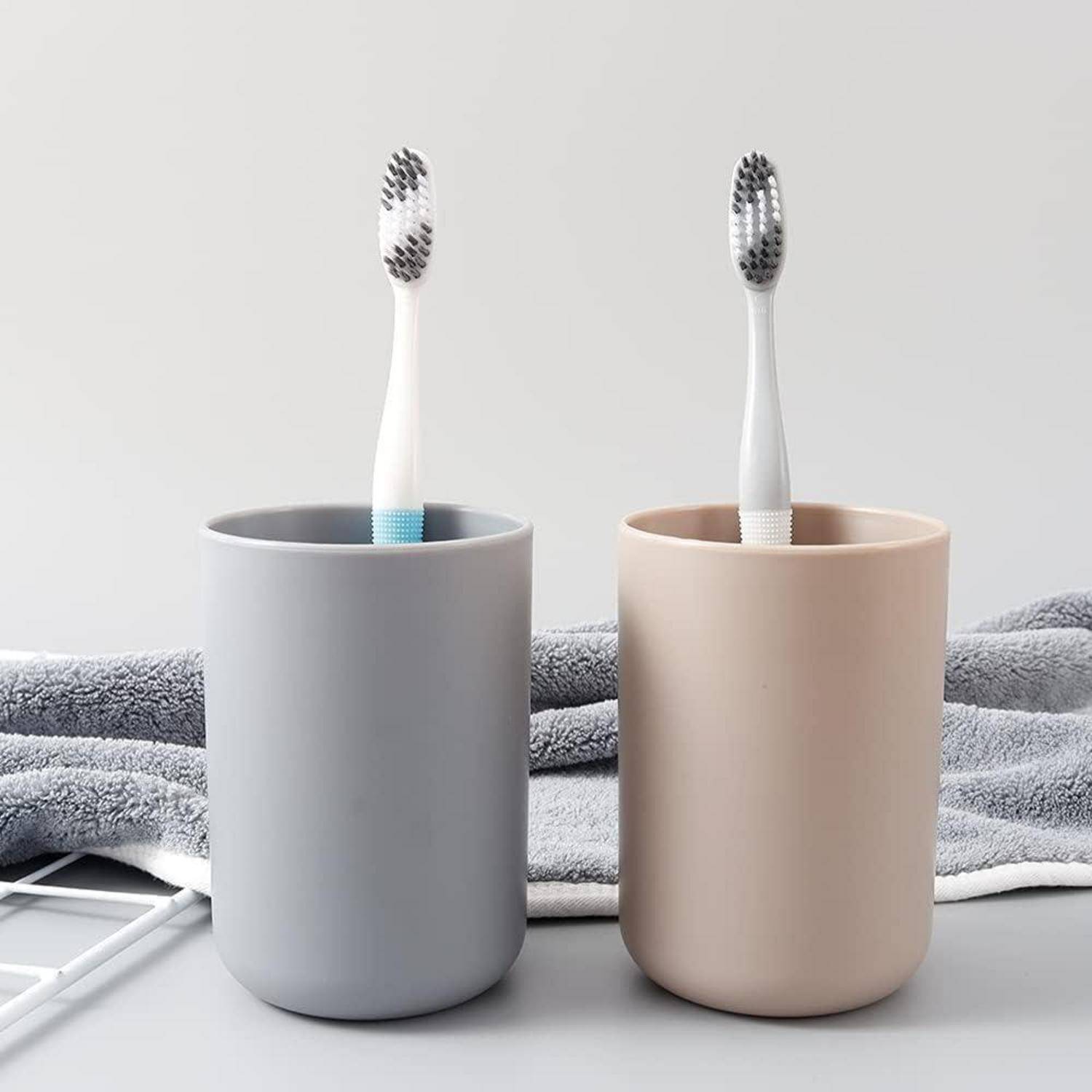 Zwei Zahnbürsten, aufrecht in separaten minimalistischen Bechern stehend, grau und beige, auf einer Arbeitsplatte mit gefalteten Handtüchern dahinter.