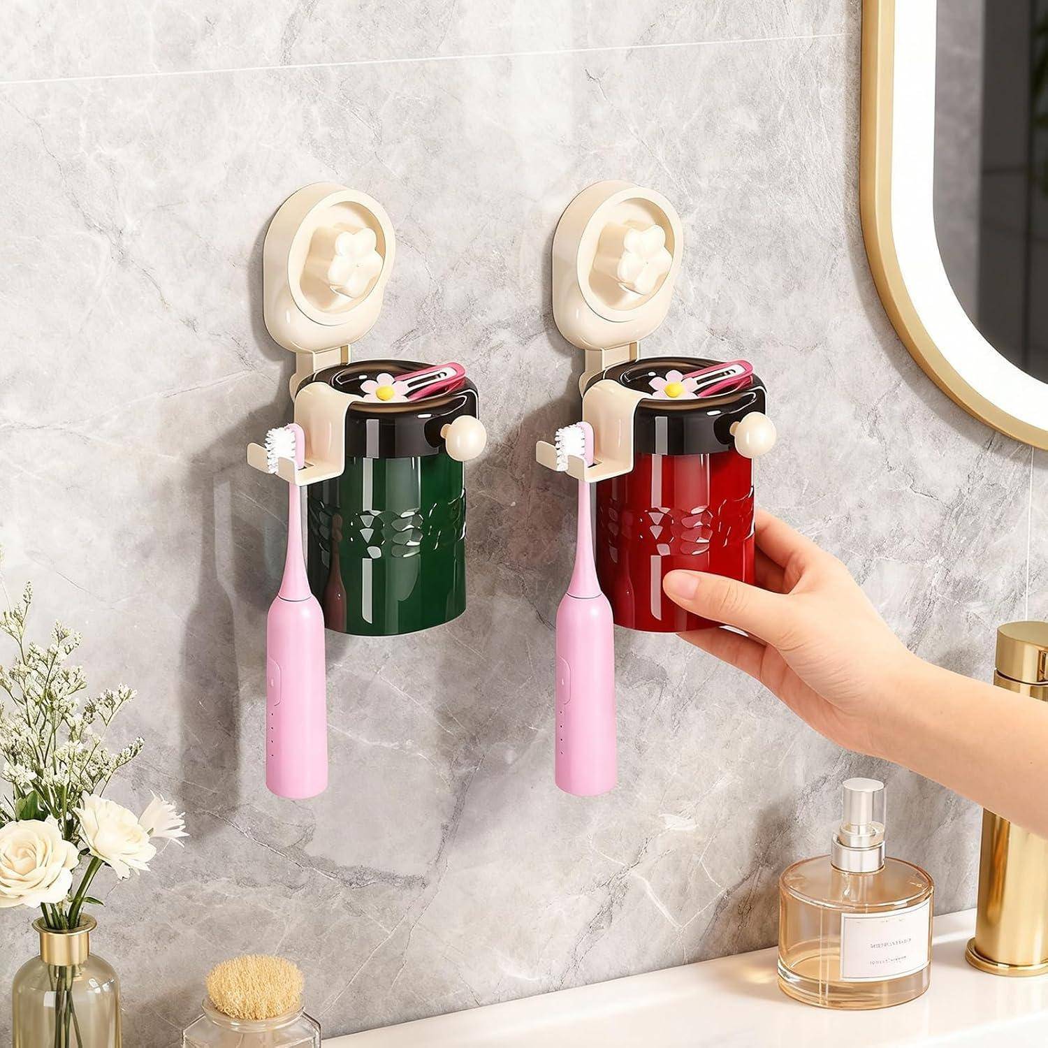 Zwei an der Wand montierte Zahnbürstenhalter mit angebrachten Bechern und Zahnbürsten, einer grün und einer rot, in einem Badezimmer; eine Hand platziert einen Becher auf dem roten Halter.