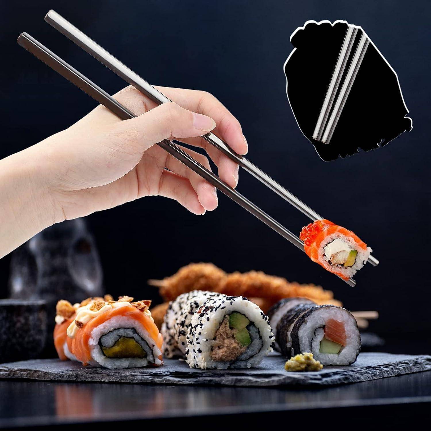 Hand hält Metallstäbchen und hebt eine Sushi-Rolle über einen Schieferstein mit verschiedenen Sushi-Stücken auf dunklem Hintergrund.