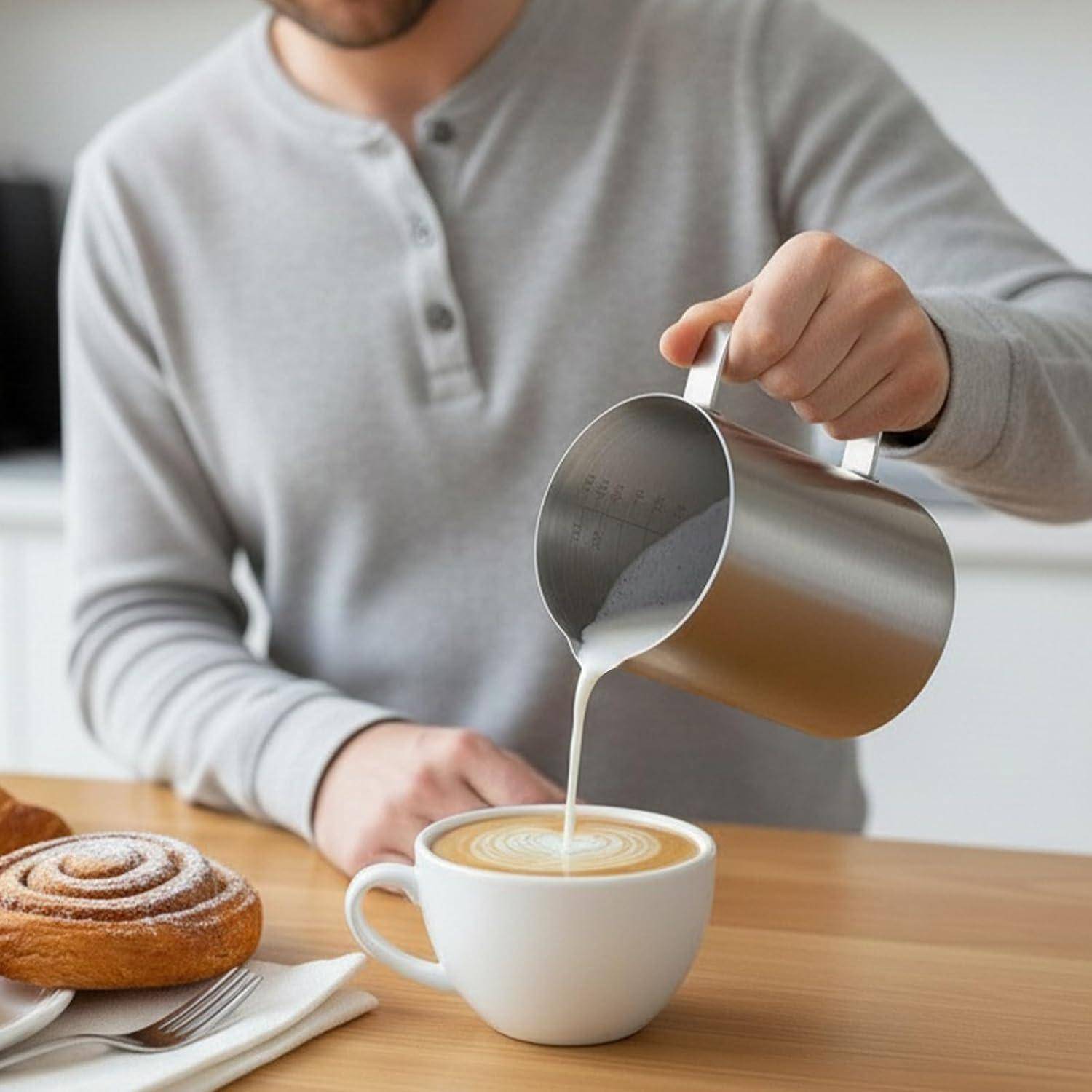 Eine Person gießt aufgeschäumte Milch aus einem Metallkännchen in einen Latte in einer weißen Tasse neben einem Gebäckstück auf einem Tisch.