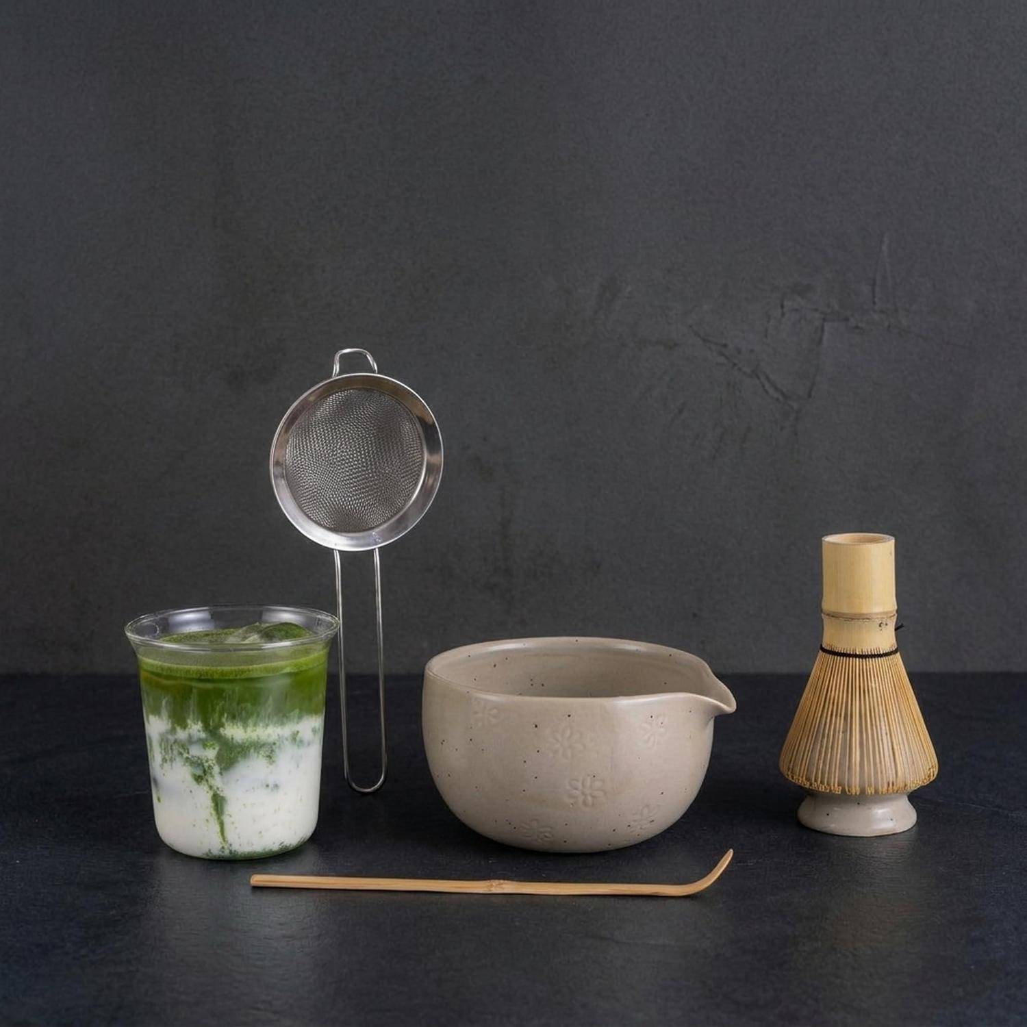 Matcha-Tee-Set mit einem Glas geschlagenem grünem Tee, einer Keramikschale, einem Sieb, einem Bamboo-Schneebesen und einem Löffel auf einer dunklen Oberfläche.