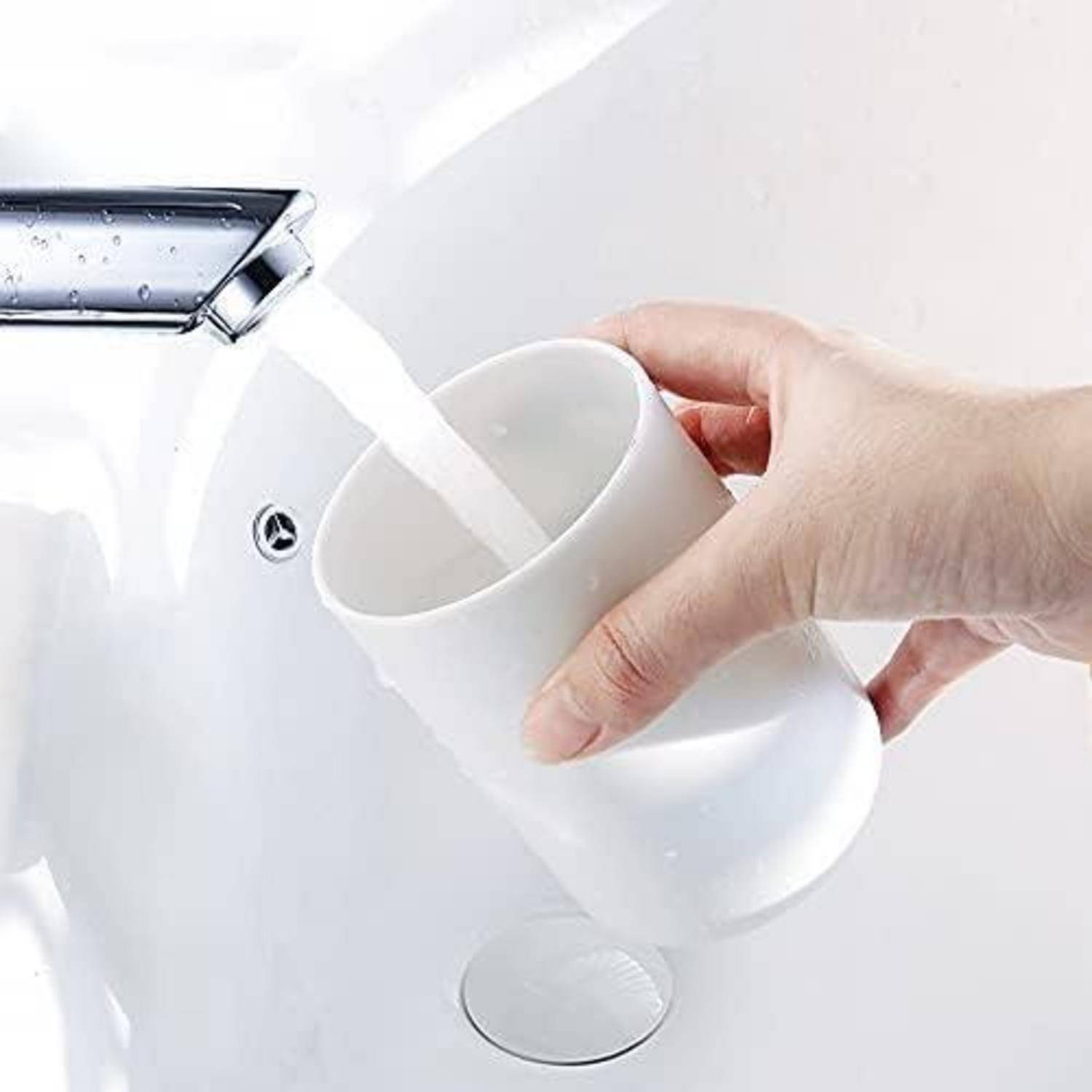 Eine Hand gießt Wasser aus einem Wasserhahn in eine weiße Tasse, die über einem Waschbeckenbecken im Badezimmer gehalten wird.