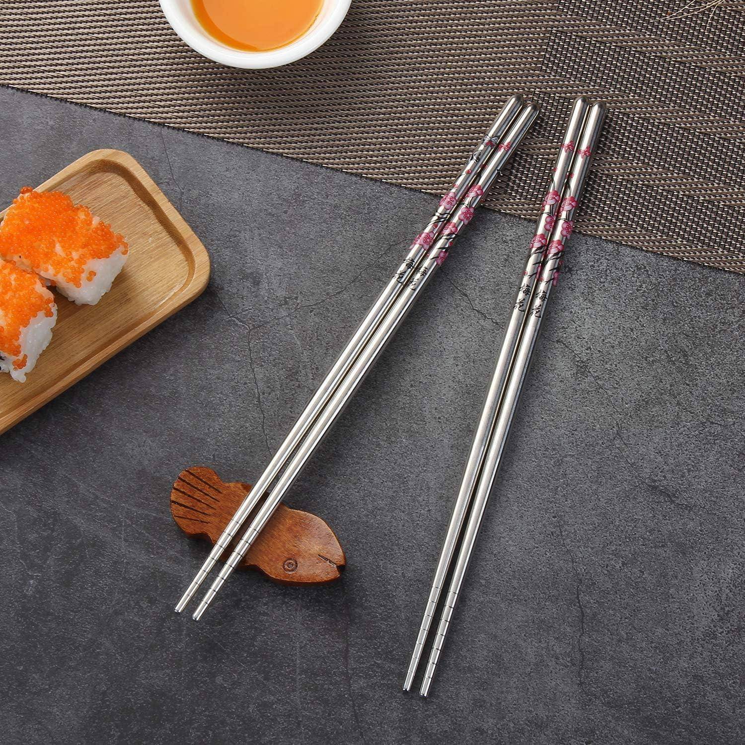 Zwei silberne Essstäbchen mit rosa Blumenverzierungen neben Sushi-Rollen, einer Schüssel mit Dip-Sauce und einer kleinen fischförmigen Ablage auf einem dunklen Tisch.