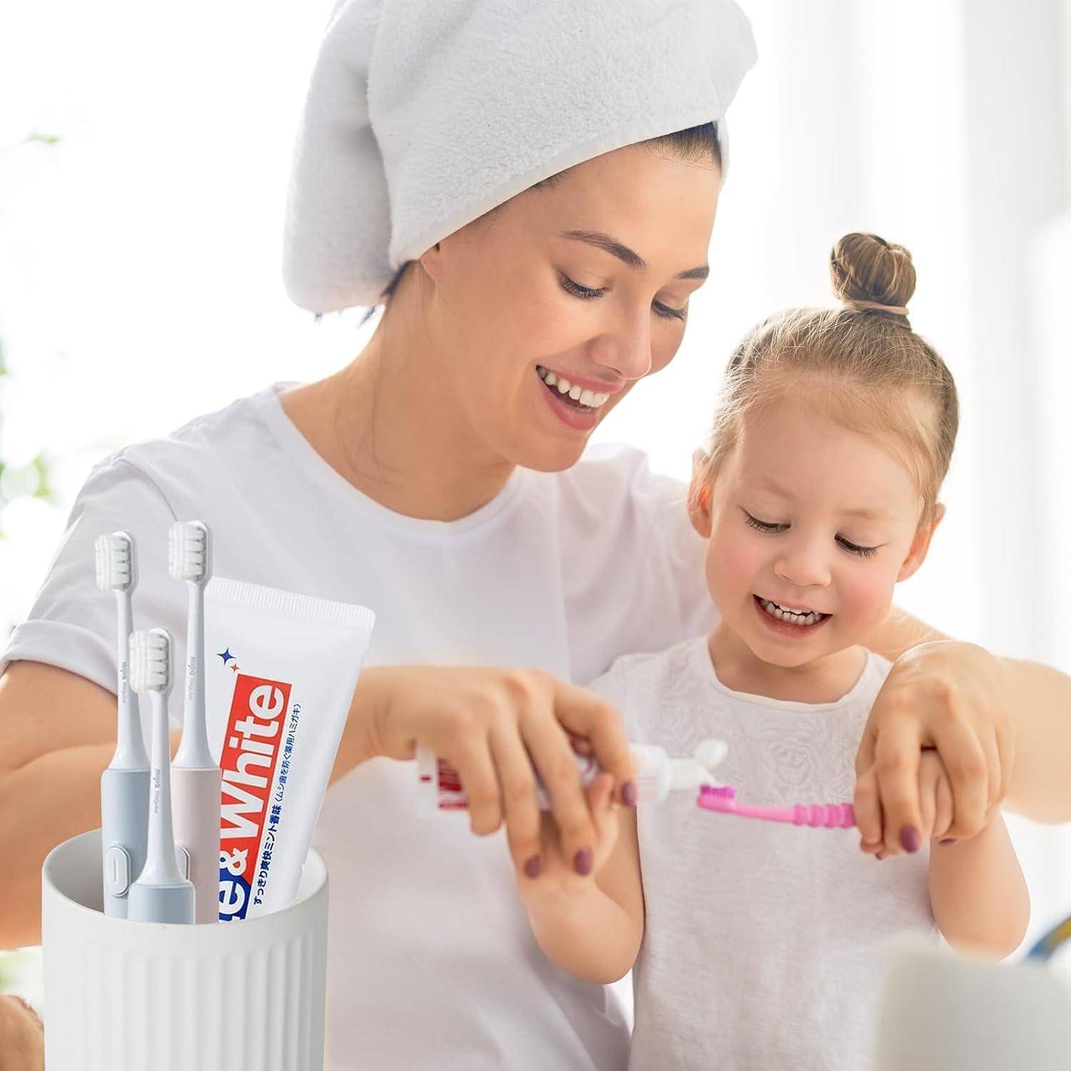 Lächelnde Mutter und kleine Tochter putzen sich zusammen die Zähne mit Zahnbürsten, neben einer Zahnpastatube und einem Zahnbürstenhalter.