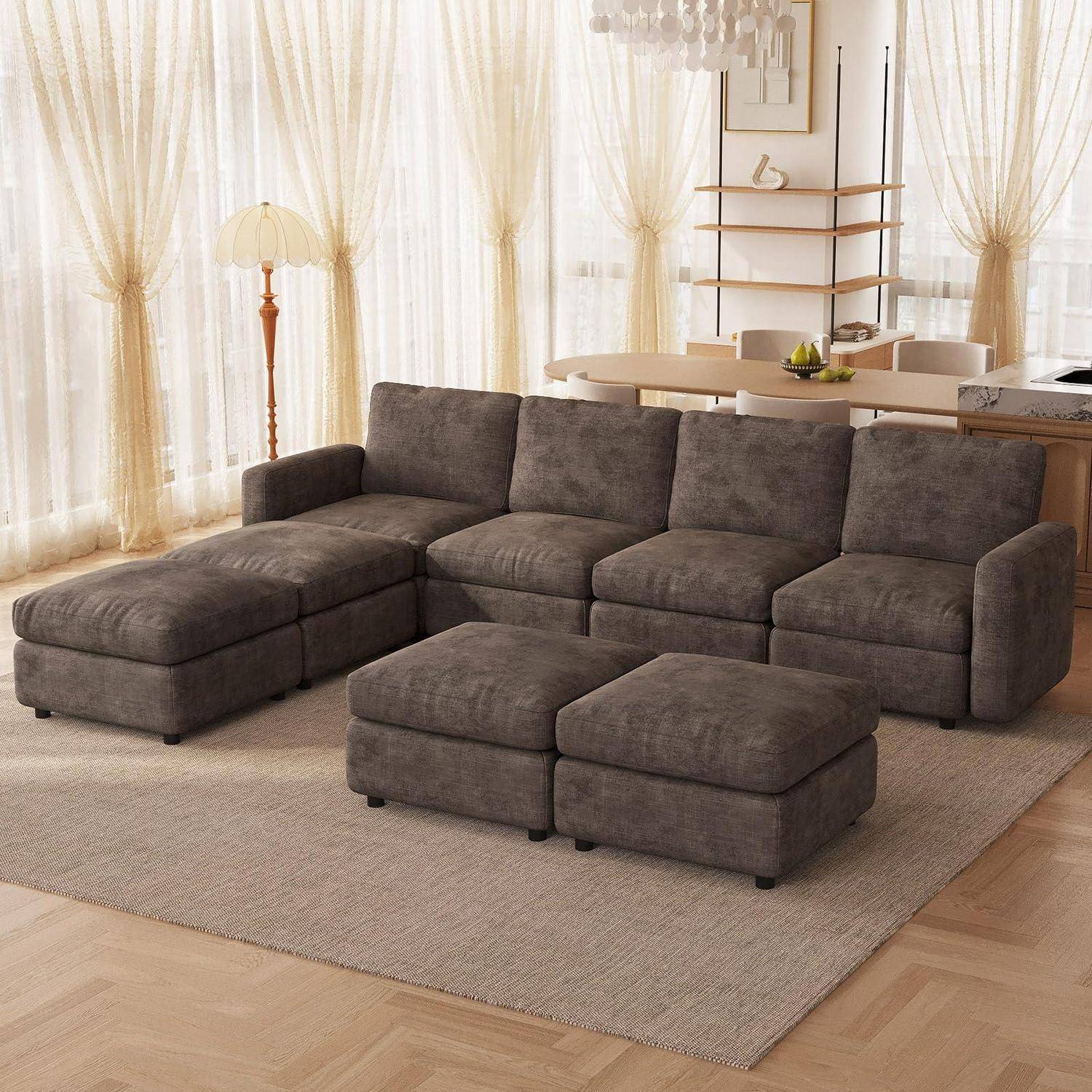 Modernes graubraunes Ecksofa mit Chaiselongue und Ottomane in einem hellen Wohnzimmer neben einem Esstisch und transparenten Vorhängen.