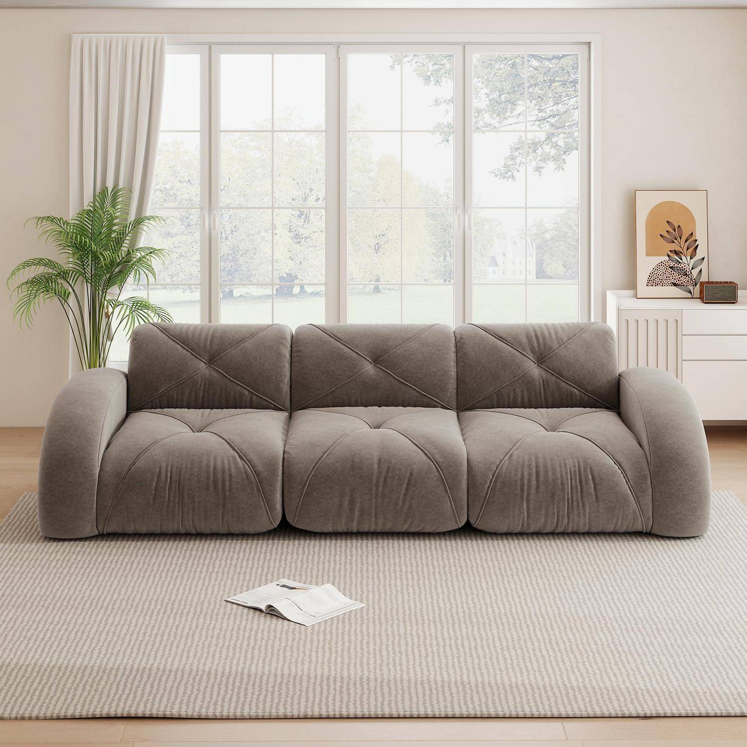 Modernes graues Dreisitzer-Sofa mit übergroßen Kissen in einem hellen Wohnzimmer, mit einem Teppich, einer Pflanze und einem Sideboard neben großen Fenstern.