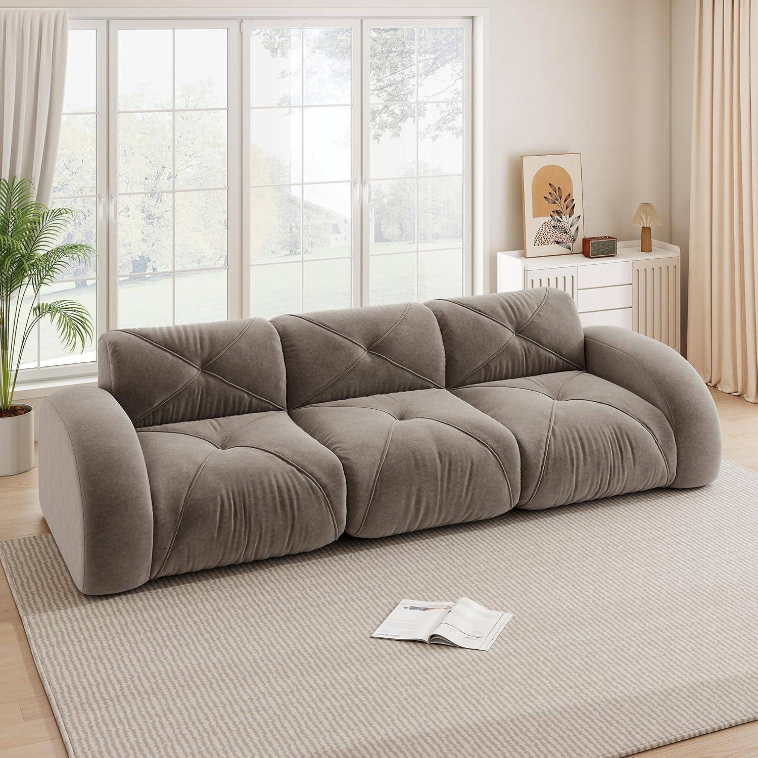 Weiches graues modulares Sofa in einem hellen Wohnzimmer mit großen Fenstern, cremefarbenem Teppich, Pflanze und Sideboard; ein aufgeschlagenes Buch liegt auf dem Boden.
