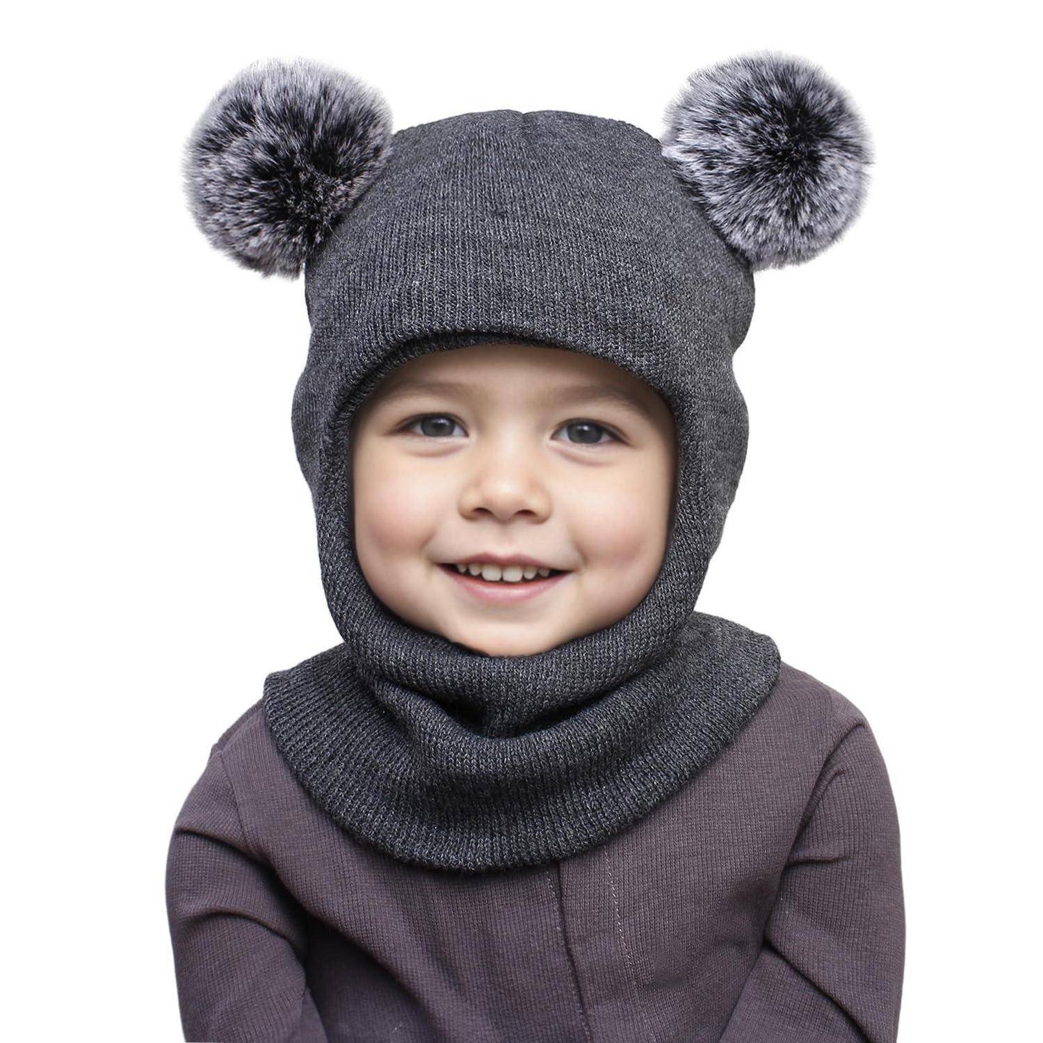 Lachendes kleines Kind, das eine graue Strickmütze mit Pompons und eine passende Gesichtsbedeckung gegen einen weißen Hintergrund trägt.