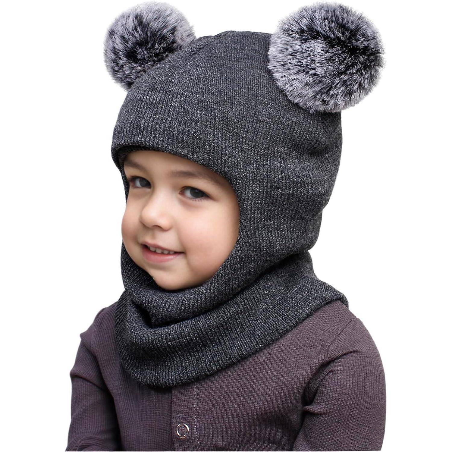 Kleines Kind, das eine graue Strickmütze mit zwei Pompons und einem passenden Halswärmer trägt, lächelt vor weißem Hintergrund.