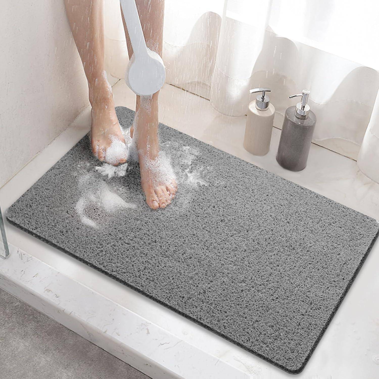 Person, die Füße auf einer grauen strukturierten Badmatte unter der Dusche wäscht, mit Seife und zwei Pumpflaschen in der Nähe.