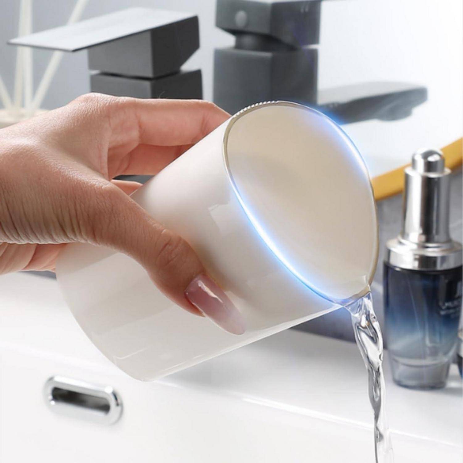 Eine Hand gießt Wasser aus einem weißen Kunststoffbecher in ein weißes Waschbecken neben einem Chromwasserhahn und einer blauen Pumpflasche.