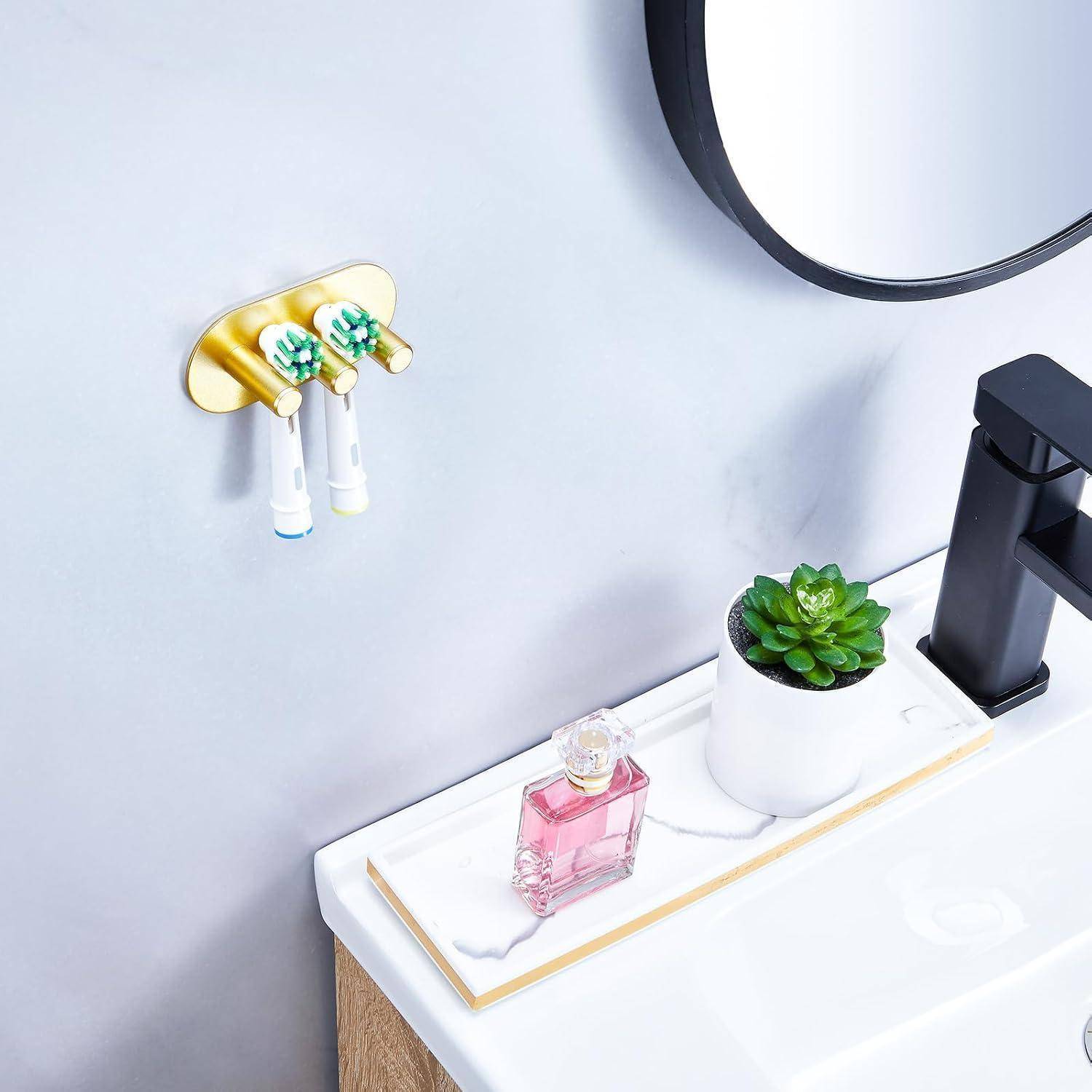 Moderner Waschbereich im Badezimmer mit an der Wand montiertem goldenem Zahnbürstenhalter, Seifenspender, Topfpflanze mit Sukkulenten, rosafarbener Parfümflasche und rundem Spiegel.